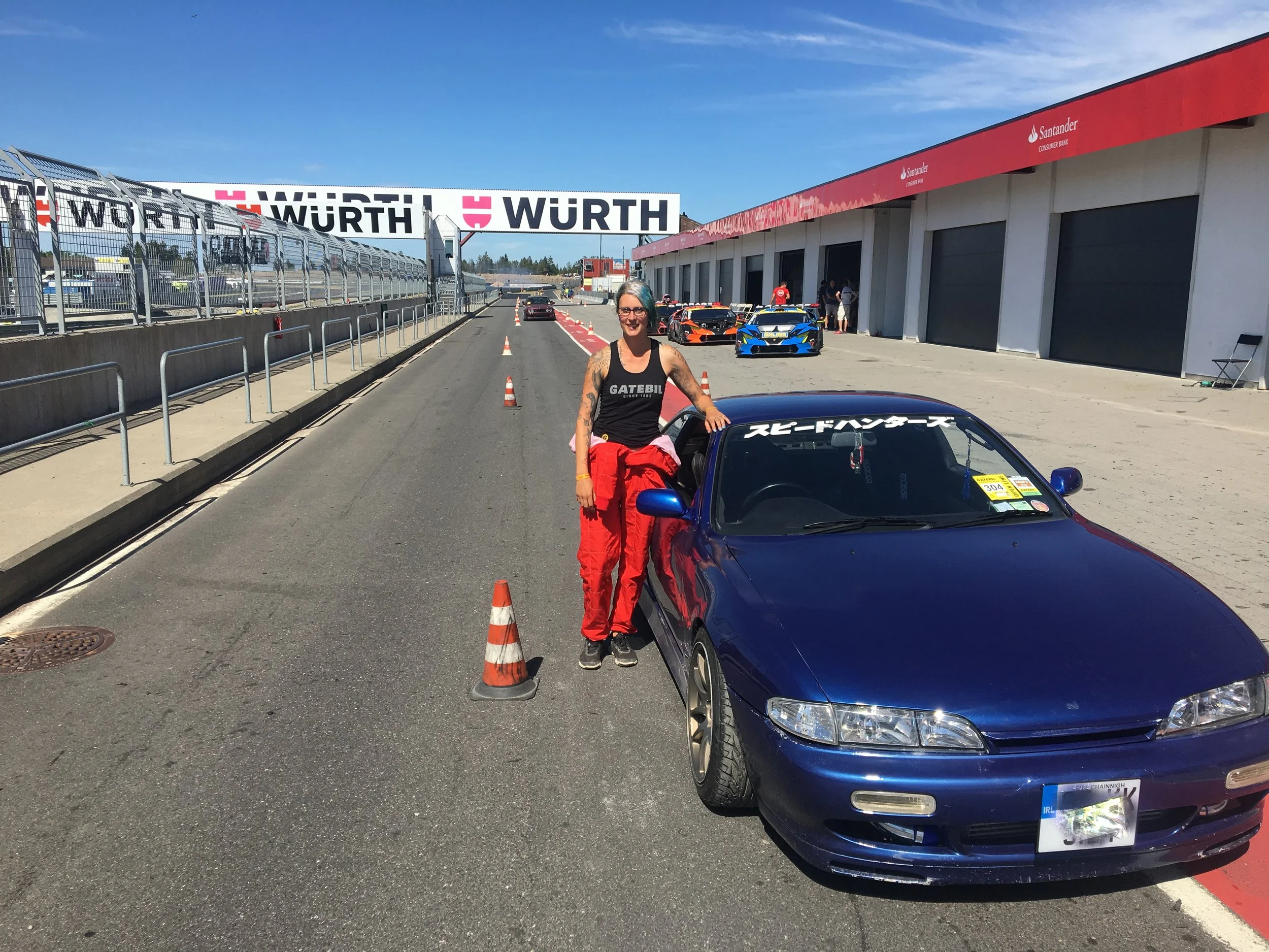 Person standing next to a blue race car on a track with signage overhead, wearing red pants and a black sleeveless shirt, surrounded by traffic cones and garages.