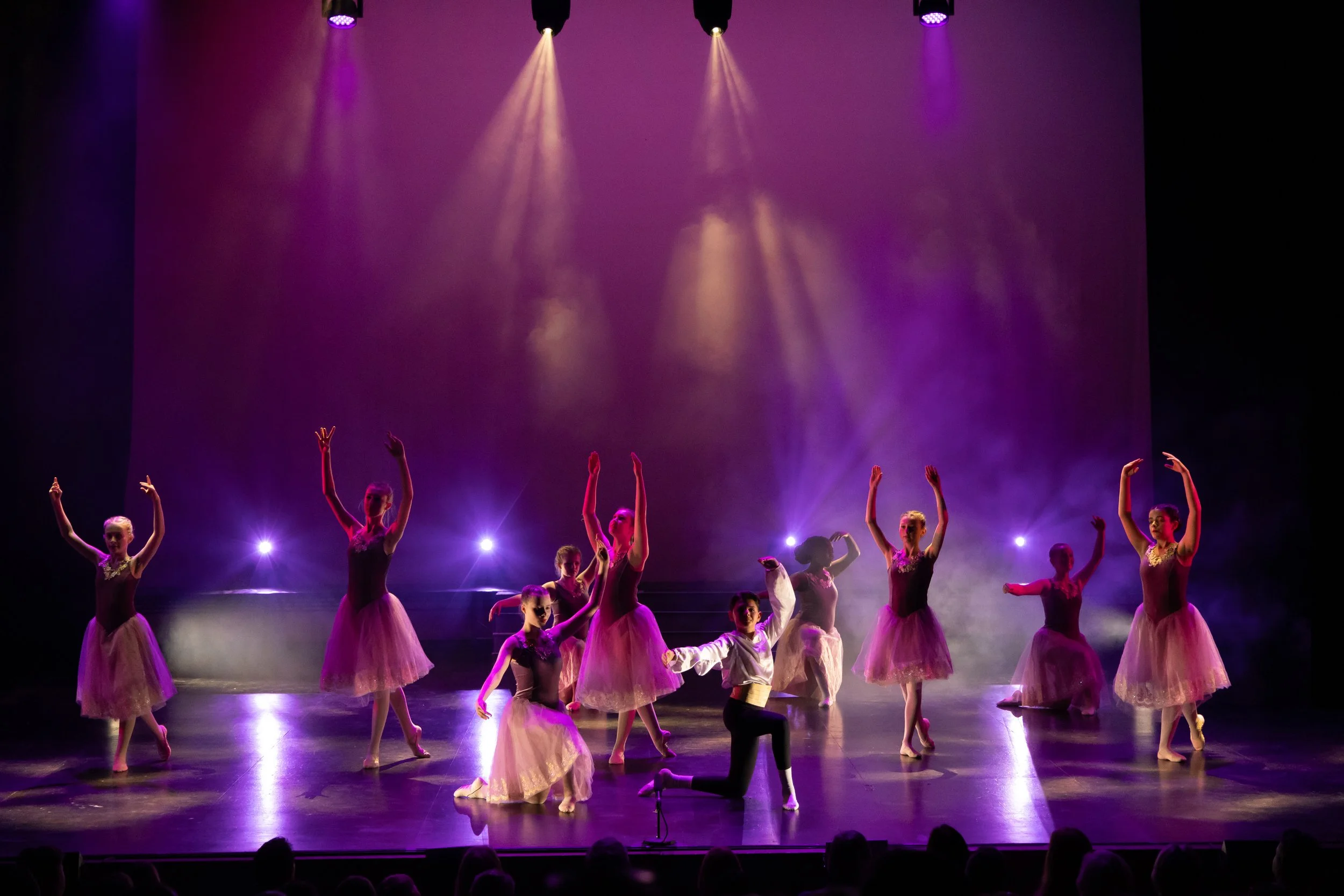 A ballet performance on stage with dancers dressed in tutus and costumes, under purple and yellow stage lighting.