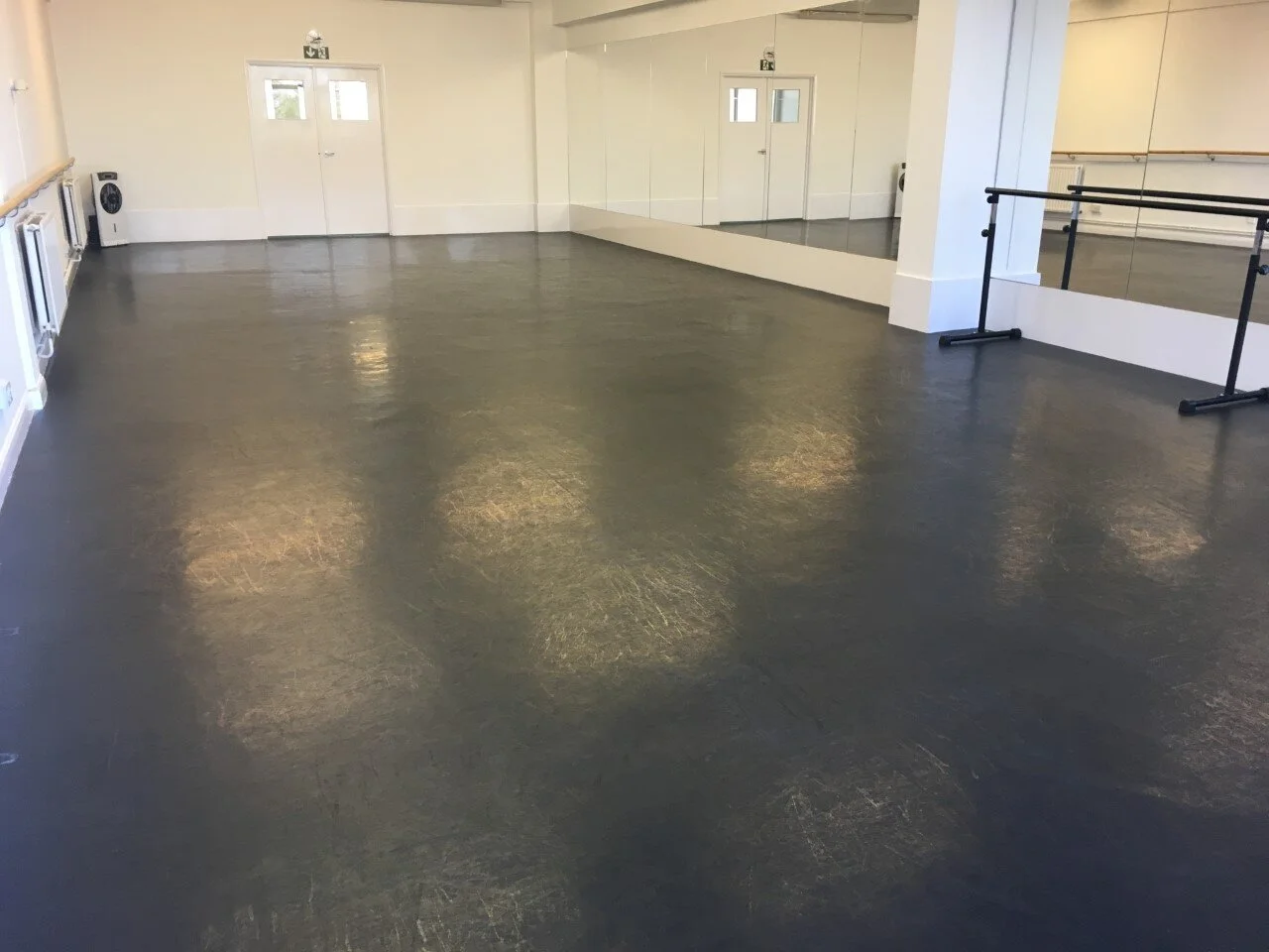 An empty dance or exercise studio with a polished black floor, white walls, mirrors along one wall, ballet barres, and doors at the back.