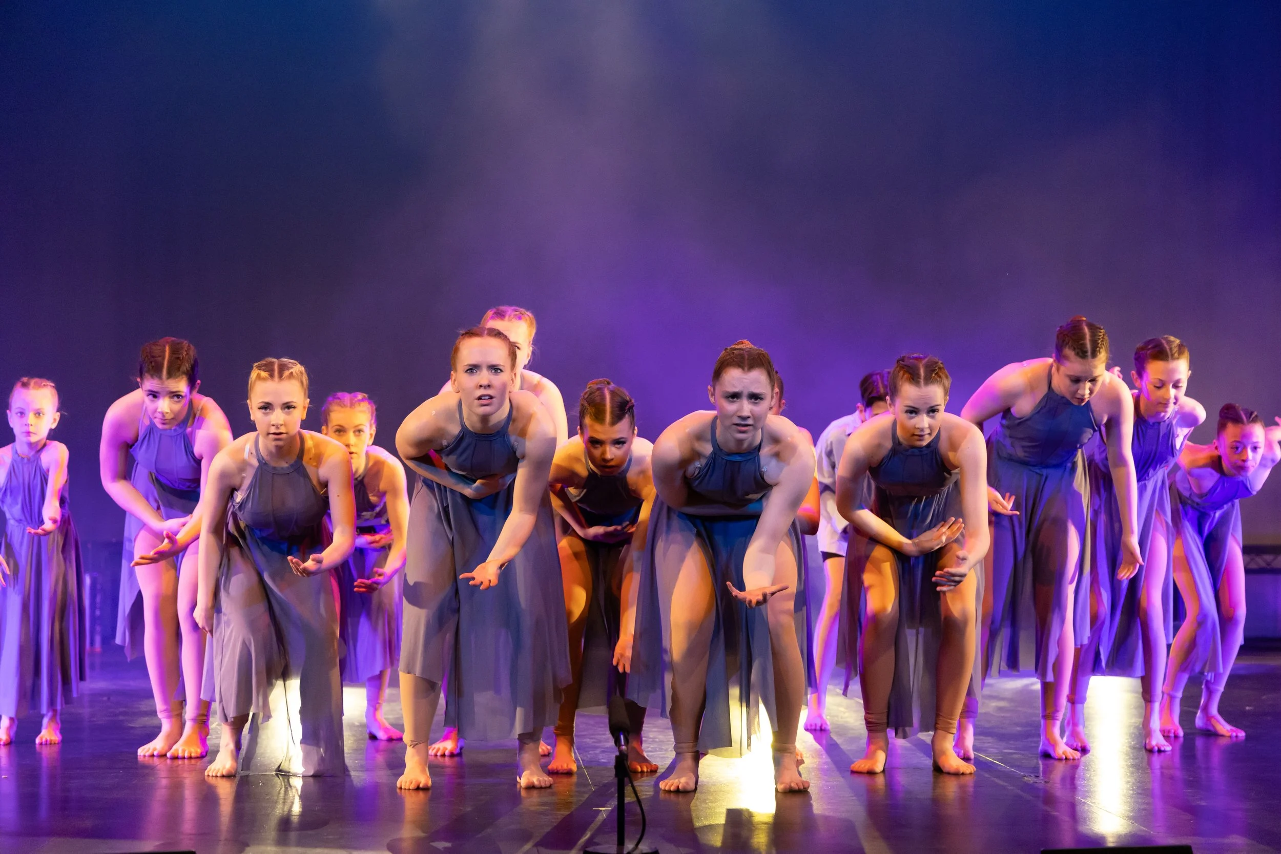 Young girls performing a dance on stage in matching purple dresses, some leaning forward with serious expressions under colorful stage lighting.
