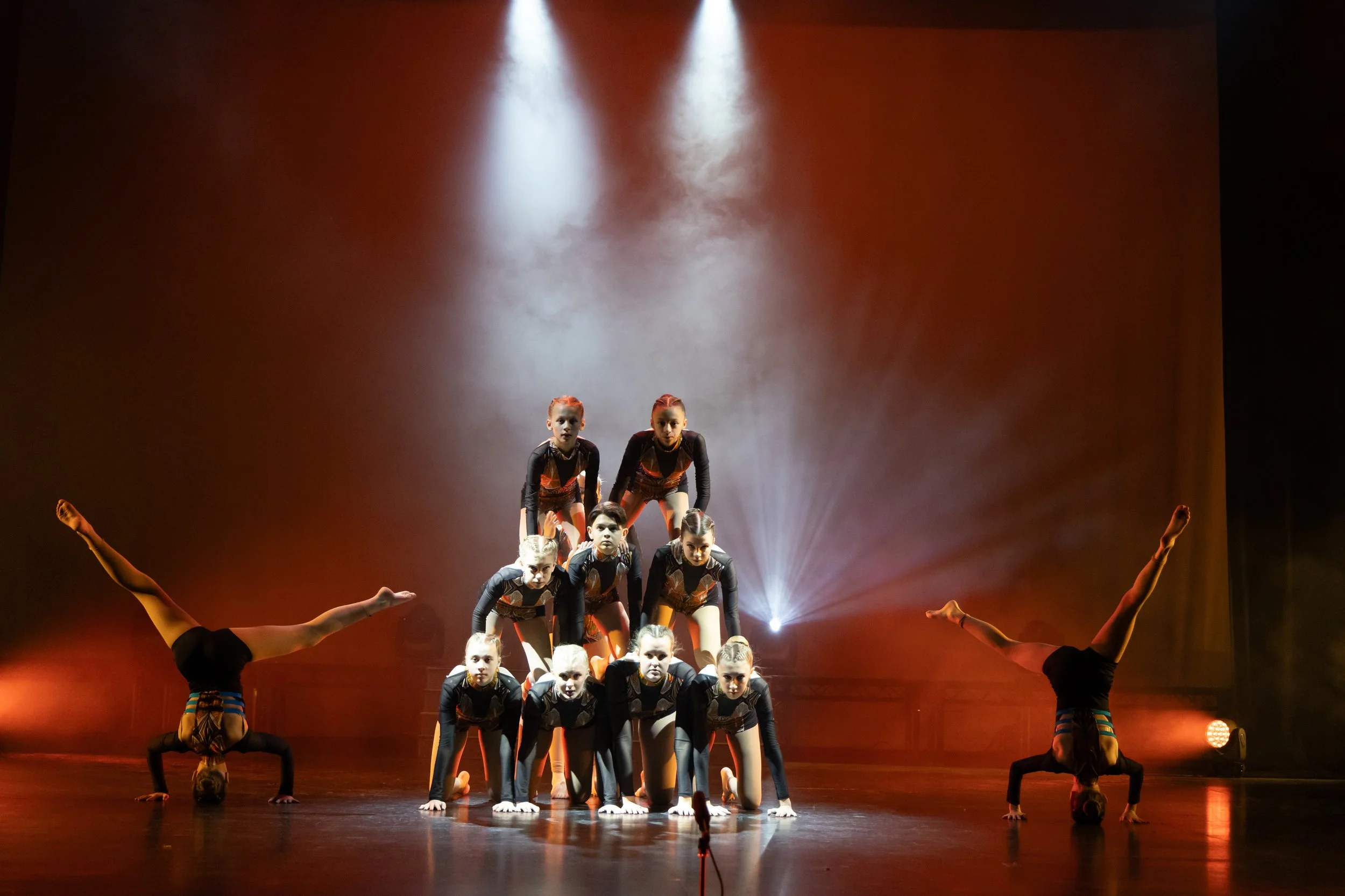 A group of young gymnasts performing a routine on stage, arranged in a pyramid formation with some doing handstands and others supporting or aiming. The stage is lit with focused spotlights and colored lighting creating a dramatic atmosphere.
