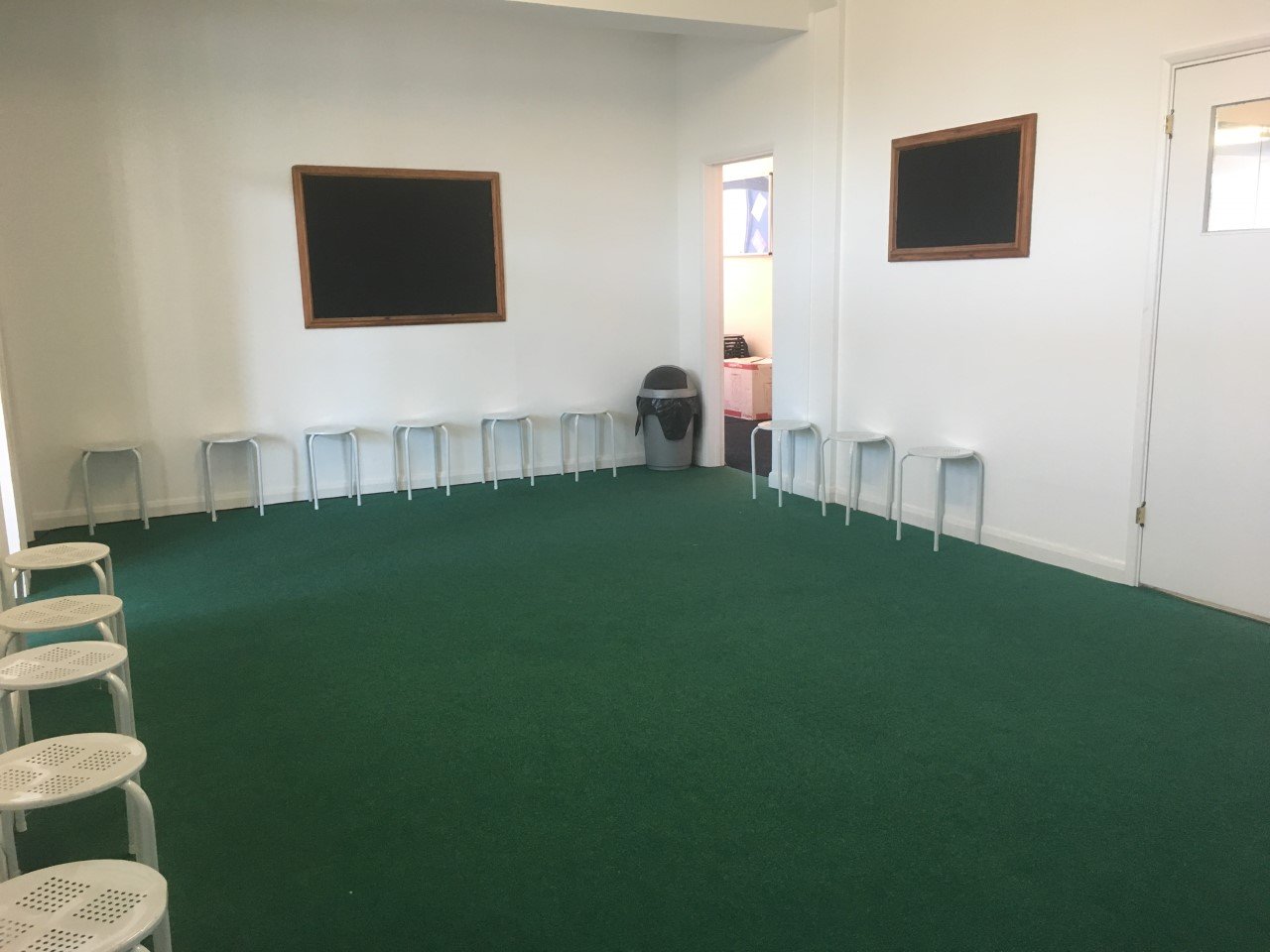 Empty room with green carpet, white walls, and small white chairs lined up along the walls. There are two small chalkboards or whiteboards framed in wood on the walls, a trash can near the door, and a small window in the door on the right side.