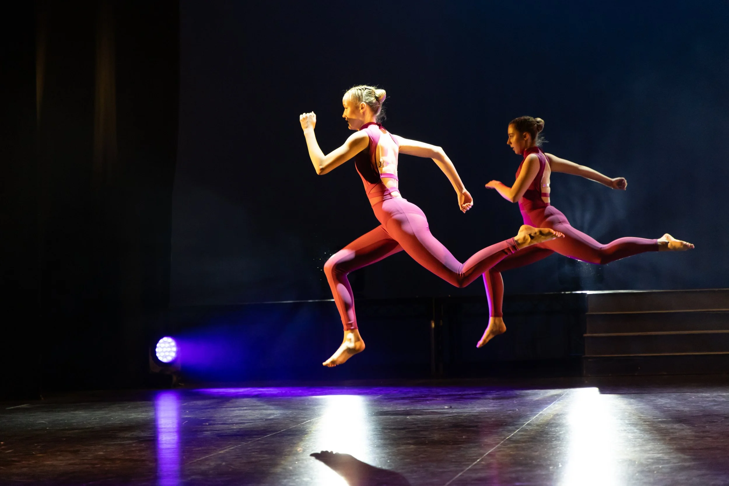 Two female dancers in pink leotards performing a leap on stage with dark background and stage lighting.