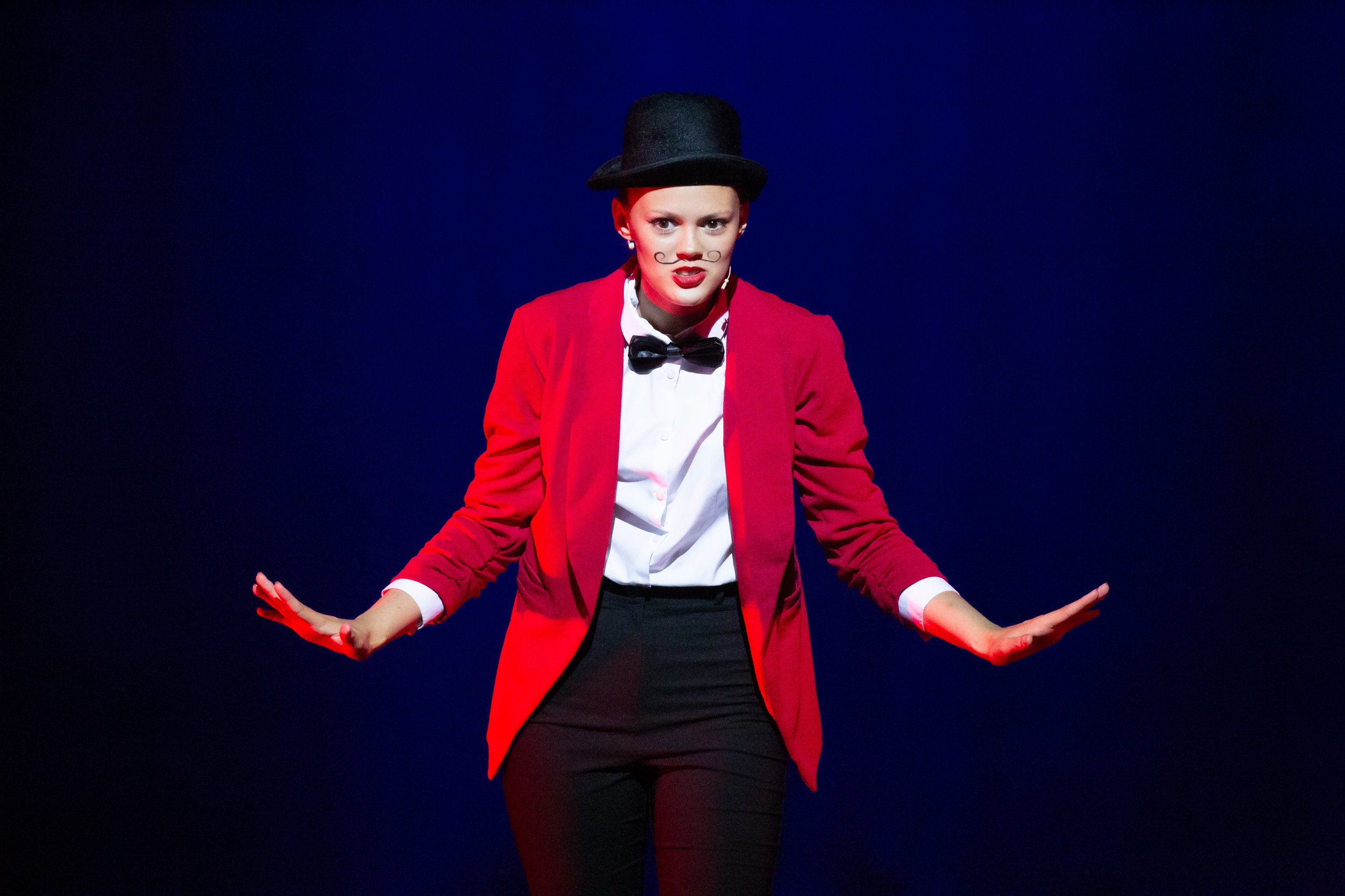 A performer dressed as a mime or clown on stage, wearing a red blazer, white shirt, black bow tie, black pants, a black top hat, and makeup with a mustache and lipstick, against a dark background.