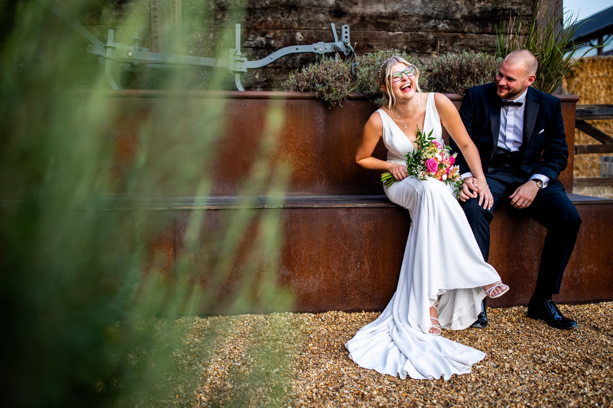 a bride and groom are sitting on a bench in front of a large wooden wall outside and the bride is holding a bouquet and wearing glasses and is laughing at the groom with a lot of fun and joy while the groom looks on with an interested look