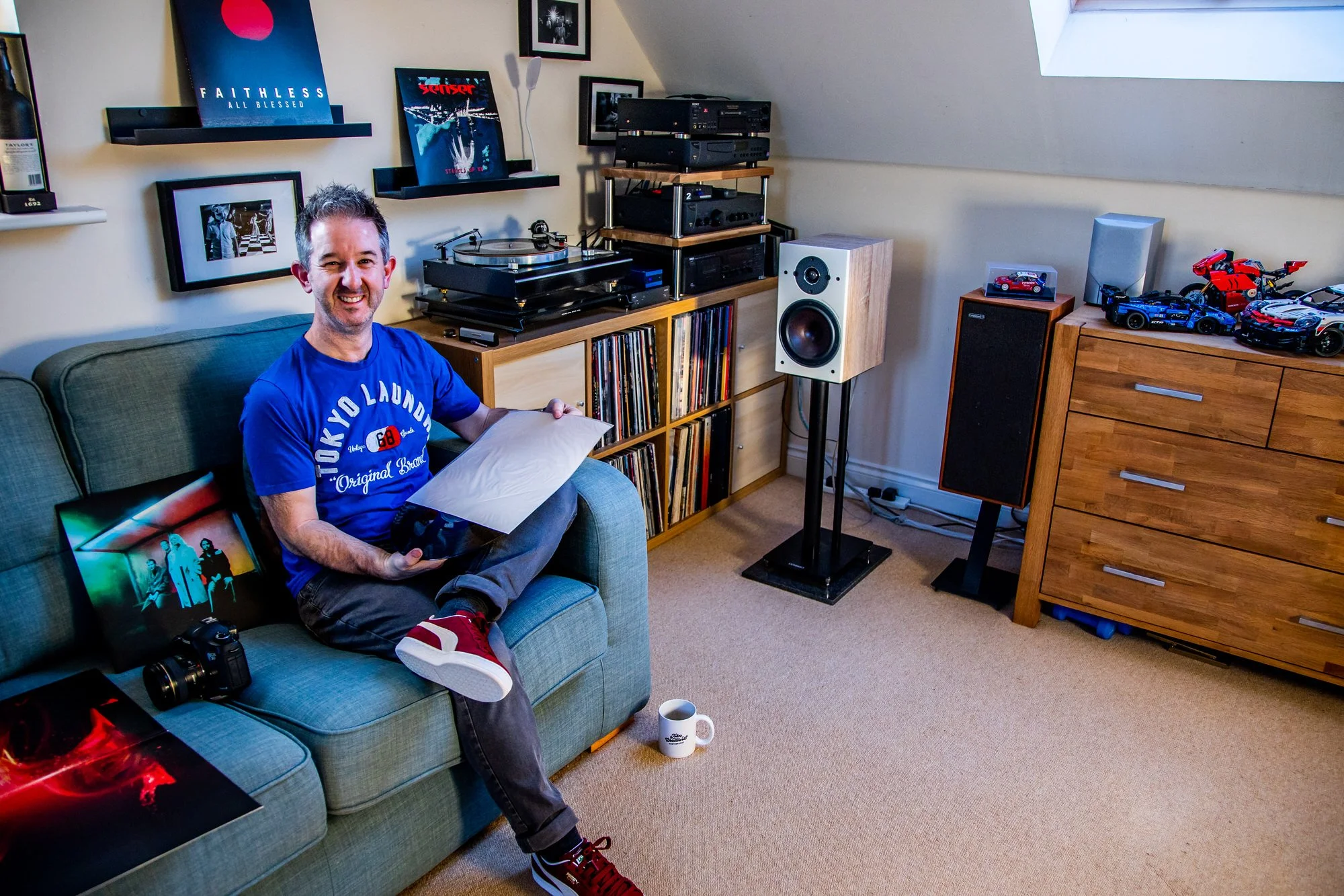 the photographer who's site this is is sitting on a sofa smiling at the camera while holding a record coming out of it's sleeve and a hifi is in the background and a camera is on the seat next to him while lots of lego is on a chest of drawers