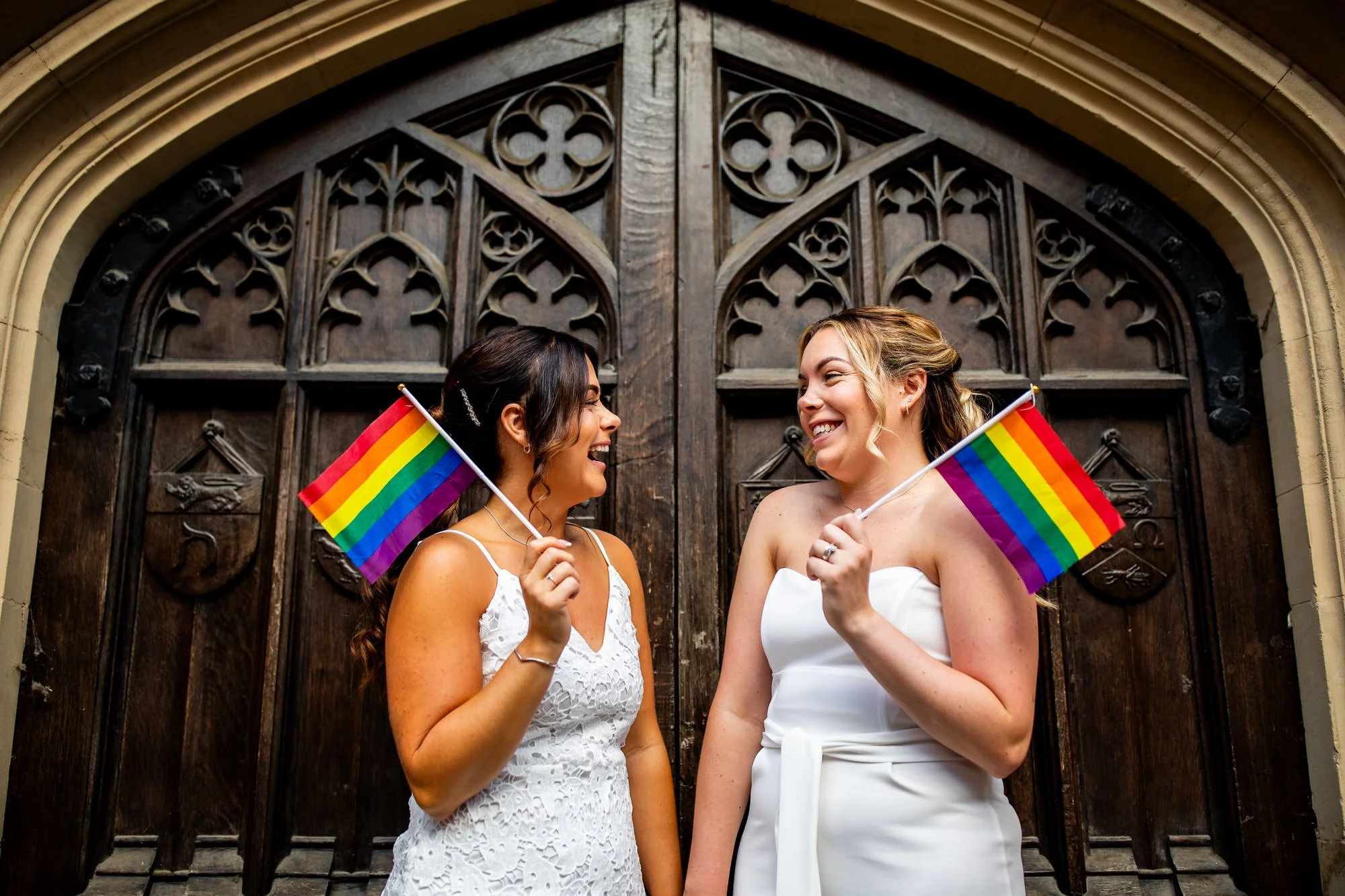 a female same sex couple who have just got married are standing next to each other in front of a large ornate wooden door and they are holding rainbow flags while looking at each other laughing and smiling