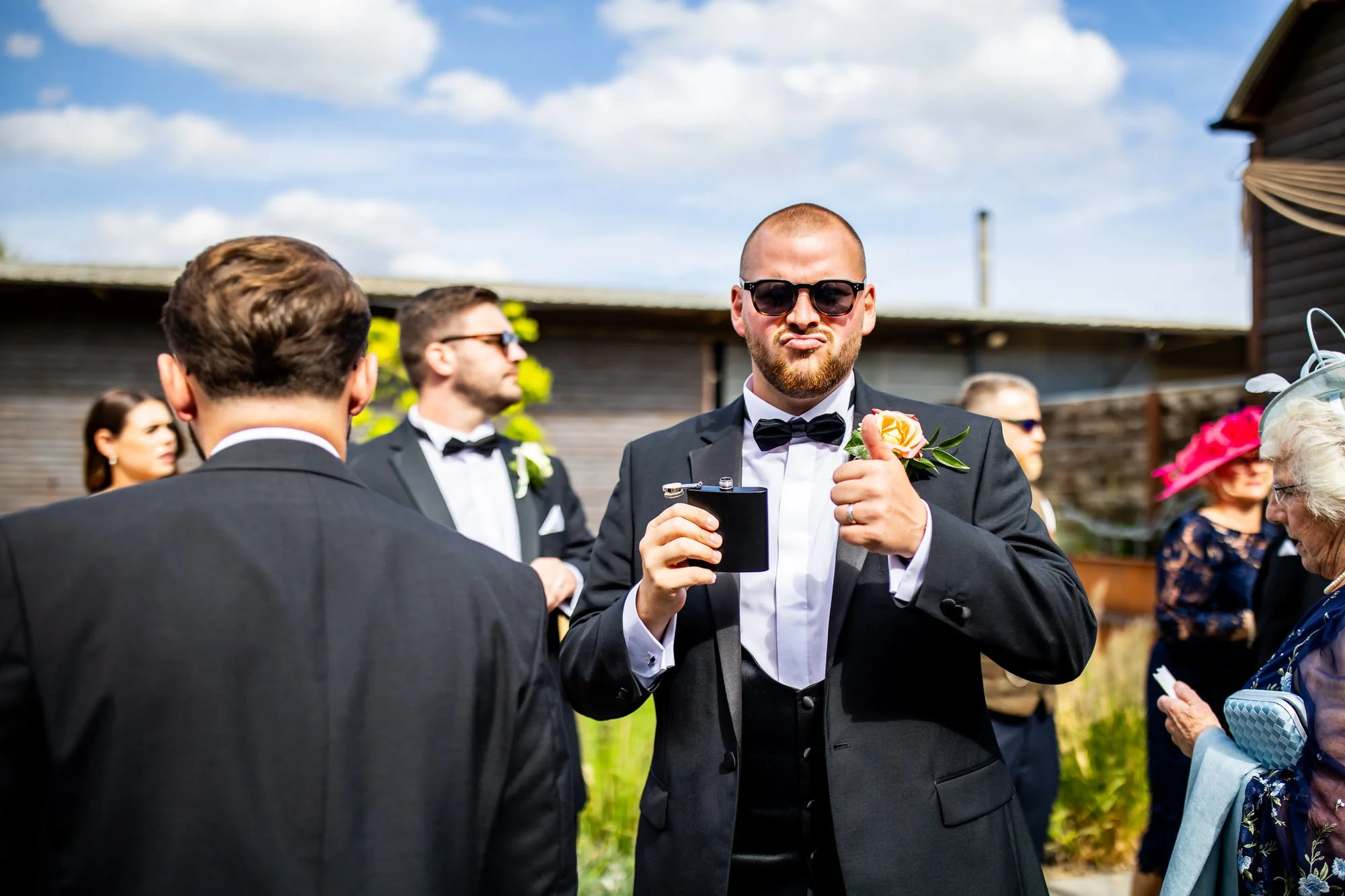 A group of people dressed in formal attire, including tuxedos and dresses, standing outdoors on a sunny day; one man in the center is wearing sunglasses, a tuxedo, holding a drink, and making a playful face.