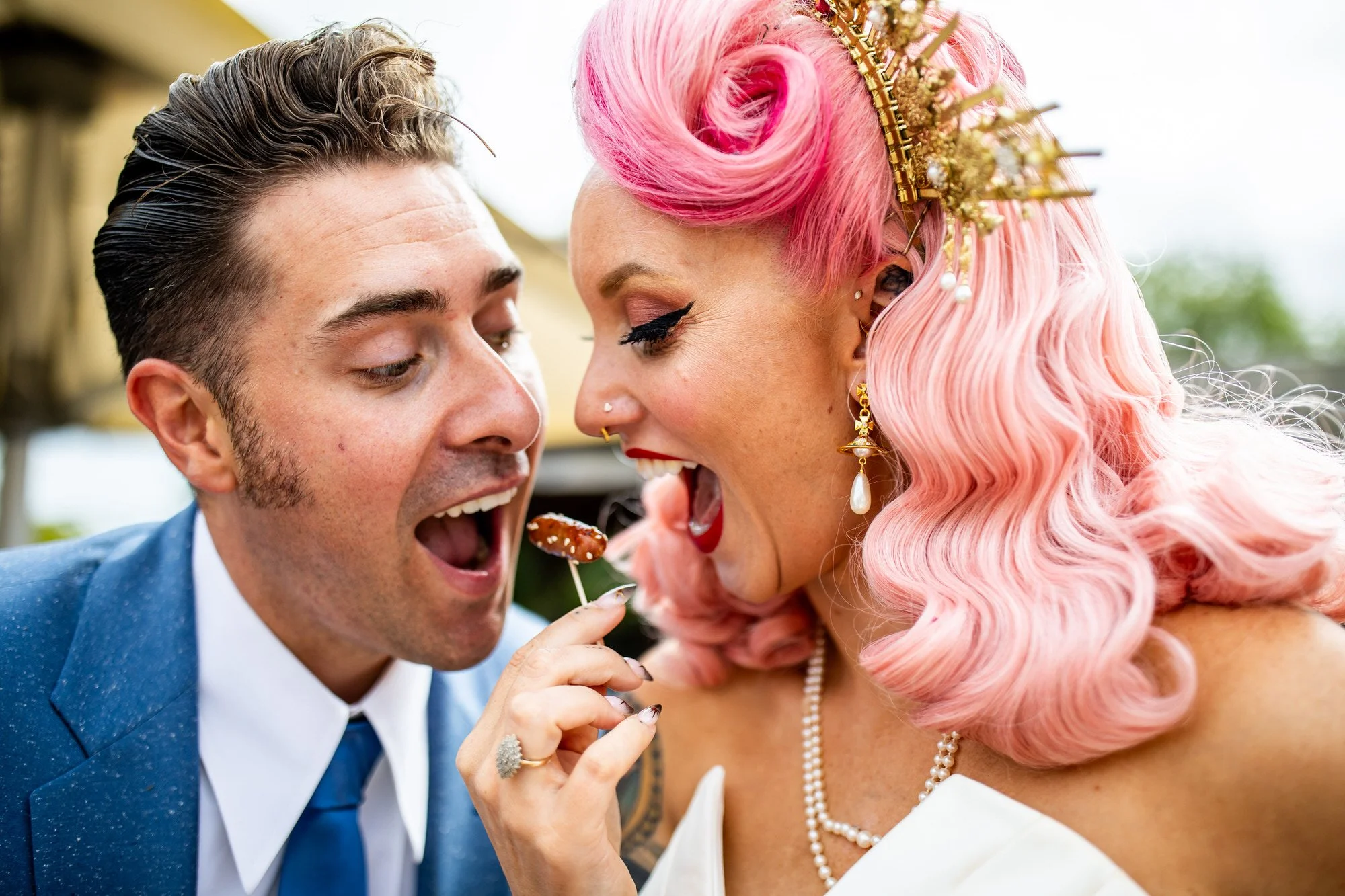 A couple dressed and styled in 50's rock and roll clothing and the bride has pink hair with a gold crown with them both pulling faces and trying to eat a sausage on a cocktail stick