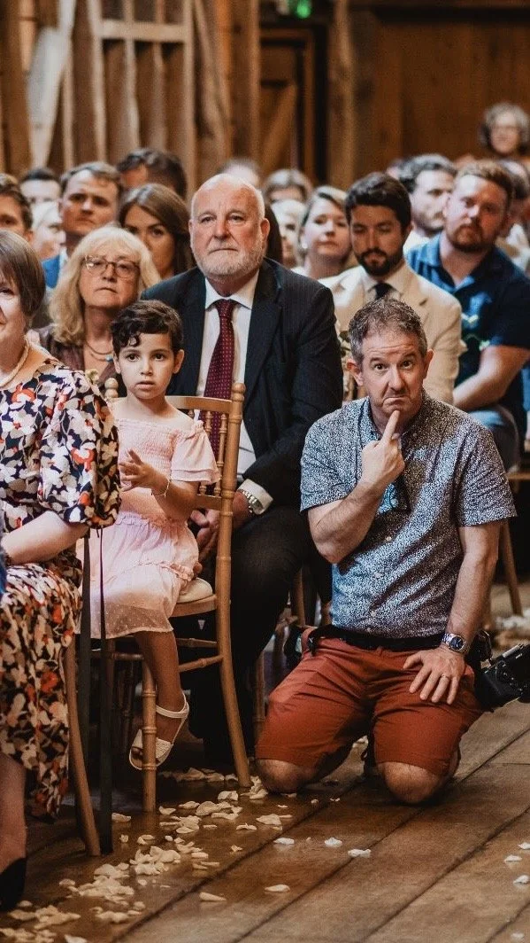 A diverse group of people seated indoors, likely at a formal event or ceremony. The image features mainly adults, with some children, sitting and attentively watching something. The setting has wooden walls and flooring with scattered flower petals on the floor.