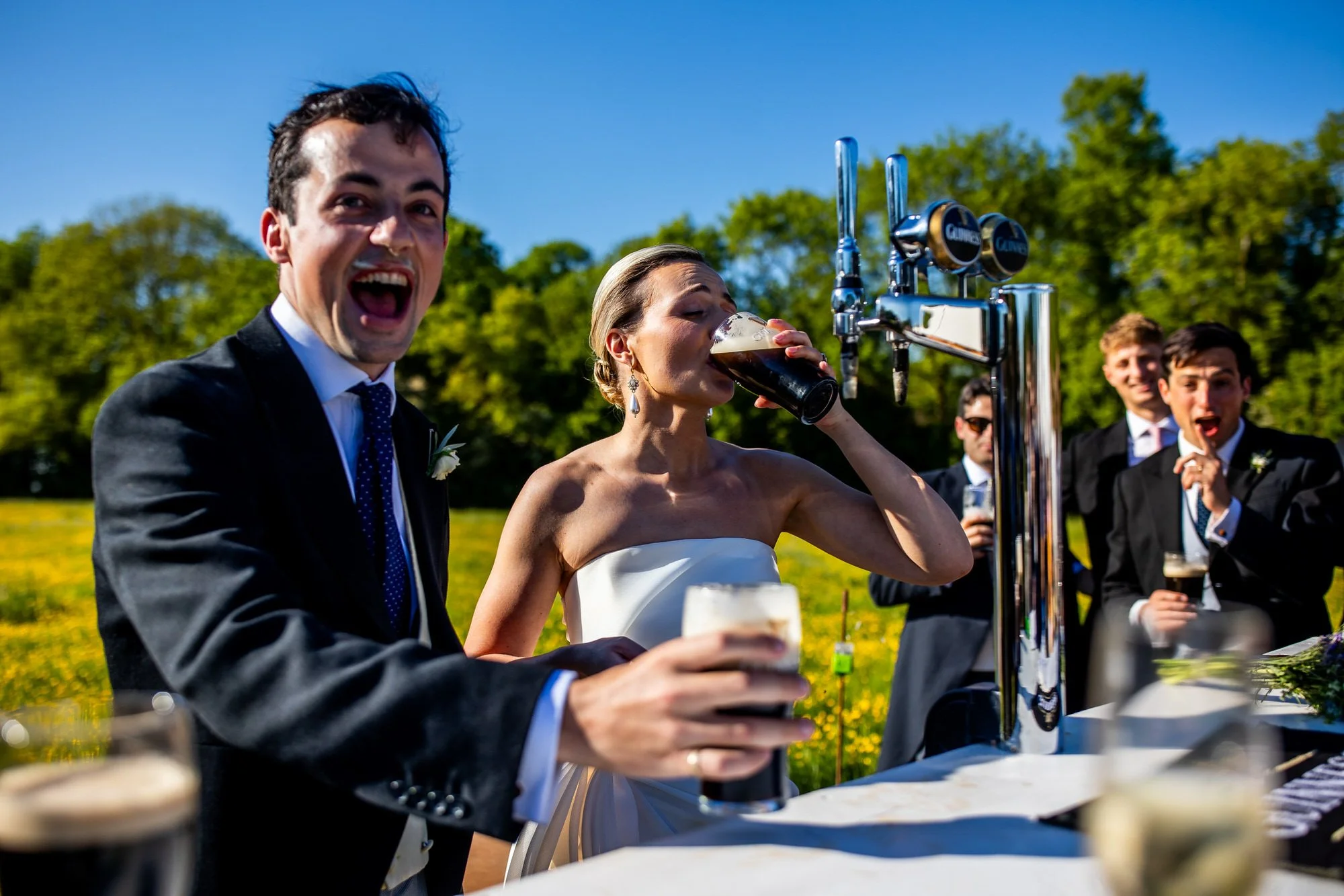 A bride in a white strapless dress and a groom in a black tuxedo celebrate outdoors with friends, one woman drinks beer from a glass, and others smile and watch in the background during daytime.
