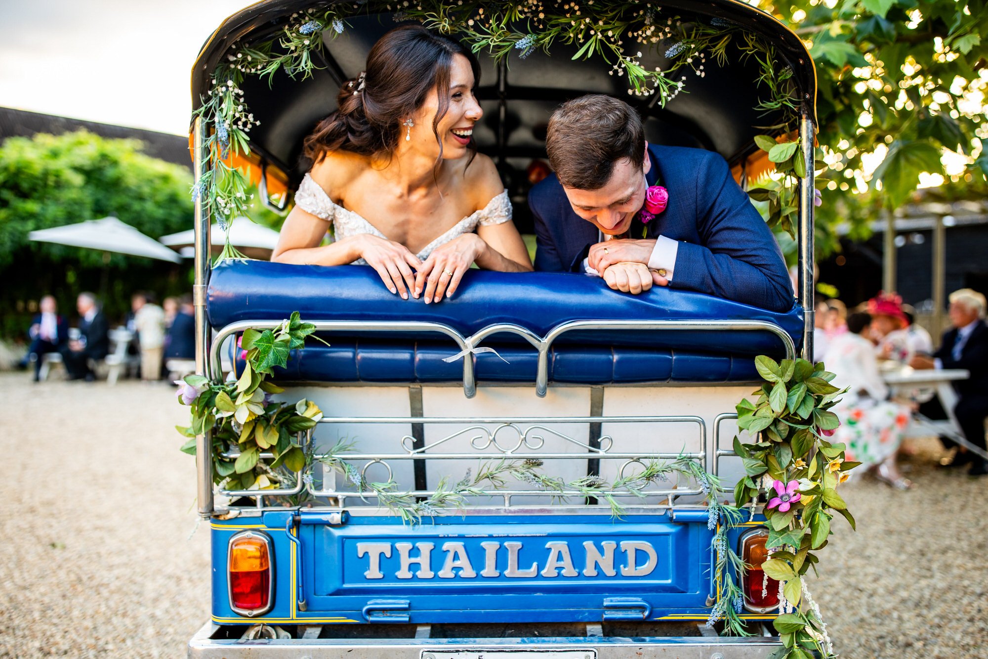 A newlywed couple in wedding attire enjoying a ride in a decorated tuk-tuk, with greenery and flowers, in Thailand during a wedding celebration.