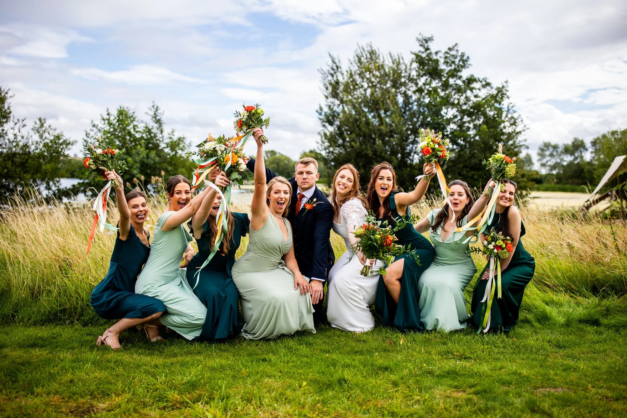 this is a group shot of the bridesmaids and the bride with the groom in the middle of them all while they are bending down holding bouquets and laughing while all on the edge of a lake at the wedding venue