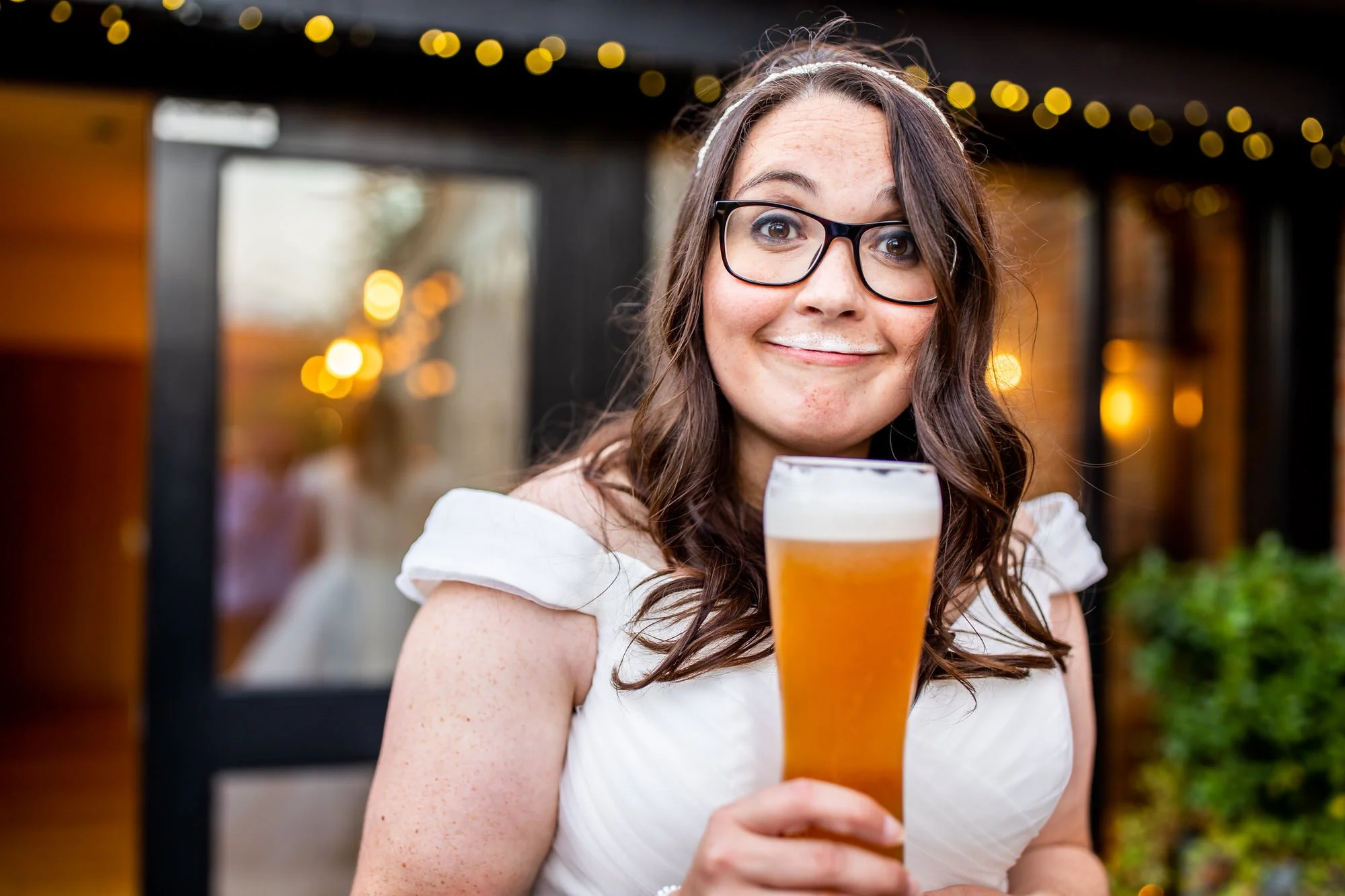 a bride is smiling funnily at the camera close up while holding a beer and has a beer froth moustache
