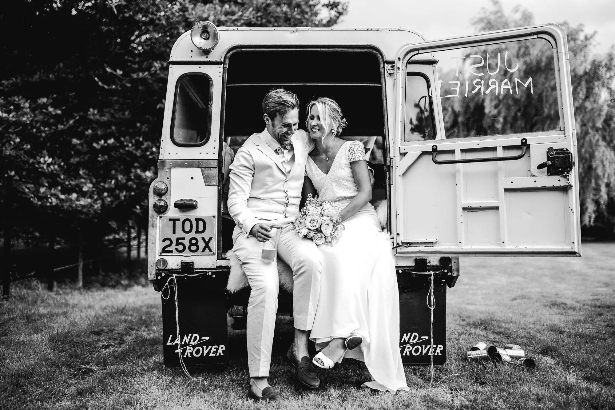 a black and white image of a couple who are sitting on the back of a land rover car with its door open and the bride is holding a bouquet and the groom is holding a beer and they are laughing at each other