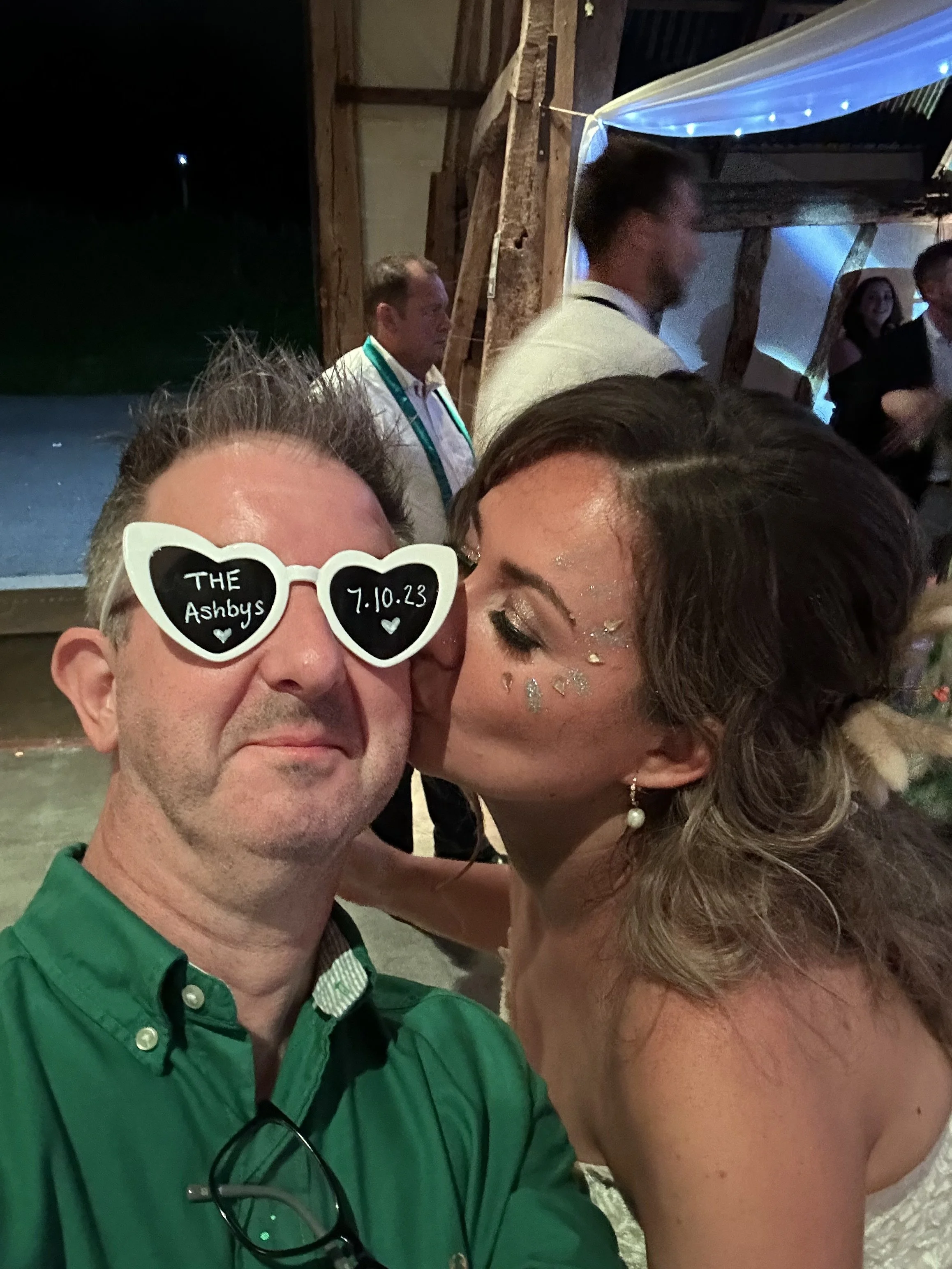 the photographer is taking a selfie while wearing a pair of white heart shaped sunglasses with the names and wedding date of the couple written on the front of them and the bride who's face is covered in glitter is kissing the photographers cheek