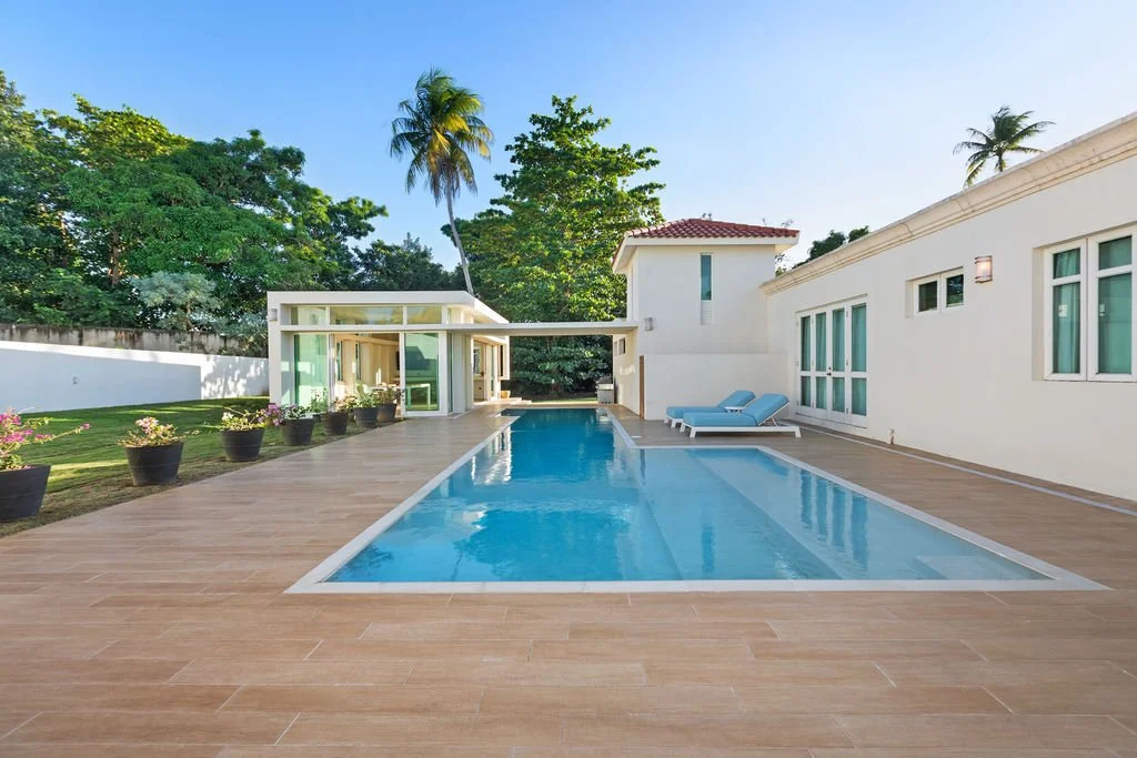 Modern backyard with a rectangular swimming pool, poolside lounge chairs, potted plants, lush trees, and a white house under a blue sky.