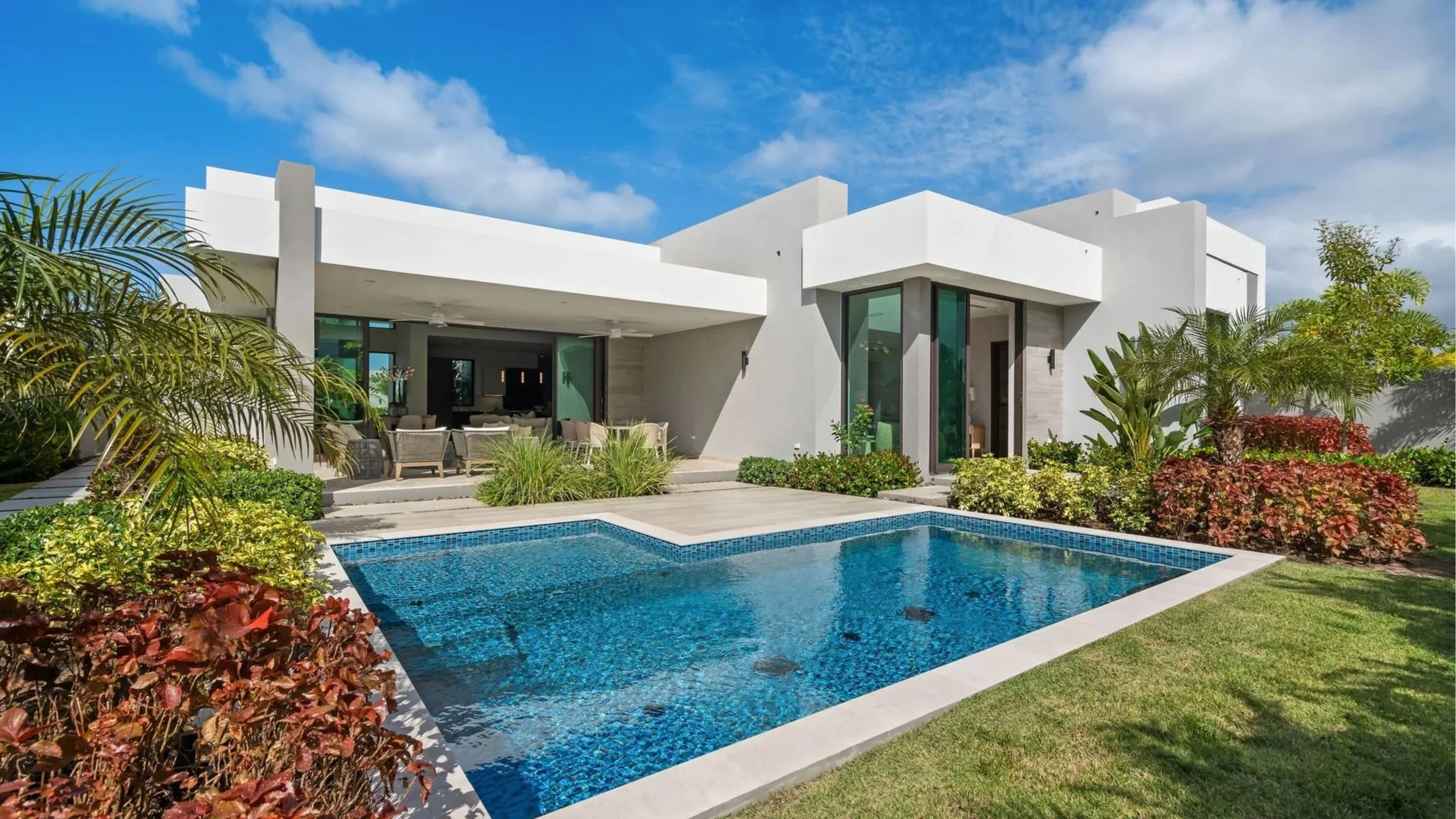 Modern white house with large glass windows and a backyard swimming pool, surrounded by lush green plants and grass under a partly cloudy sky.