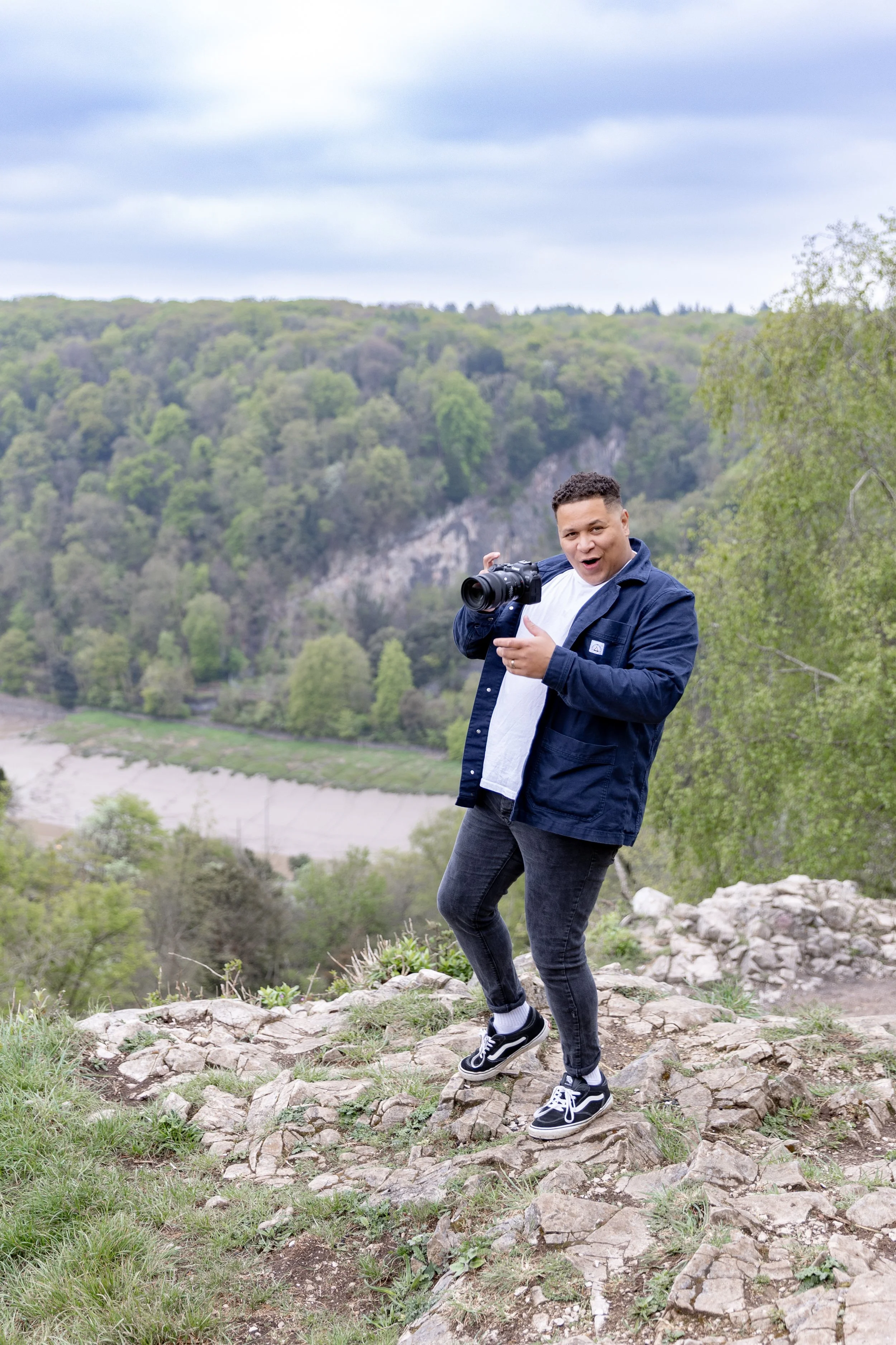 A Somerset wedding photographer in casual clothing holding a camera on a rocky outdoor trail with green trees and hills in the background.