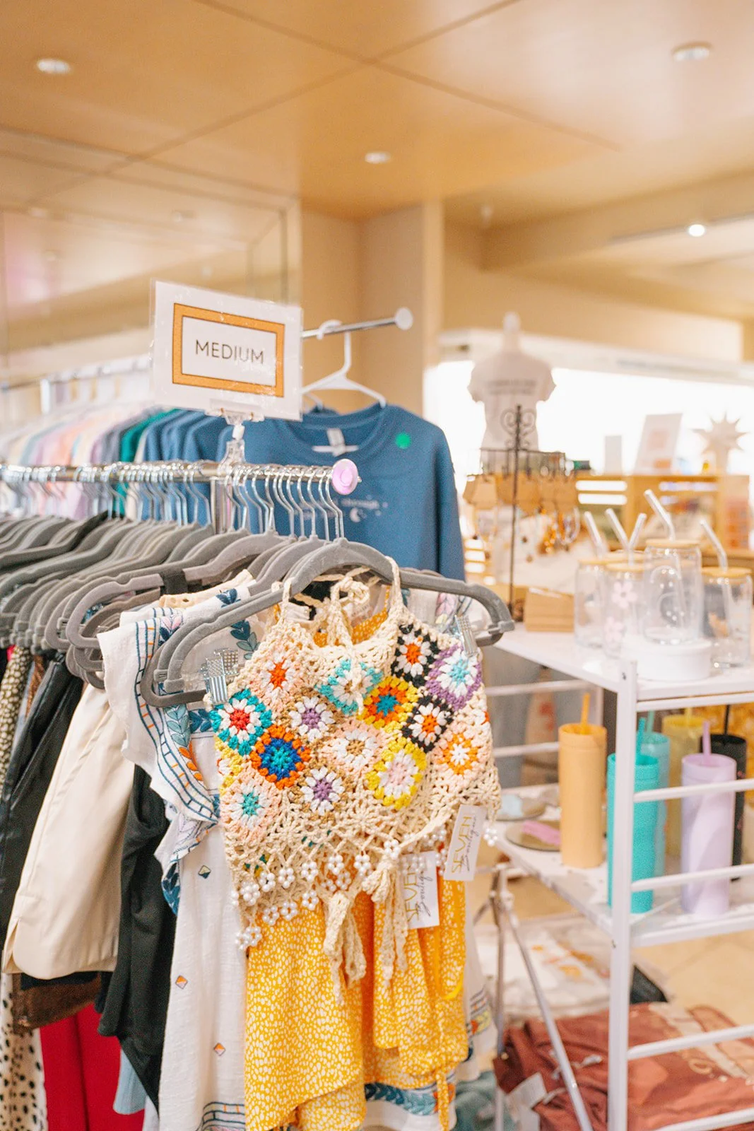 Clothing rack with colorful dresses and a crochet bag, store sign reading 'Medium', decorative items and glassware in background.