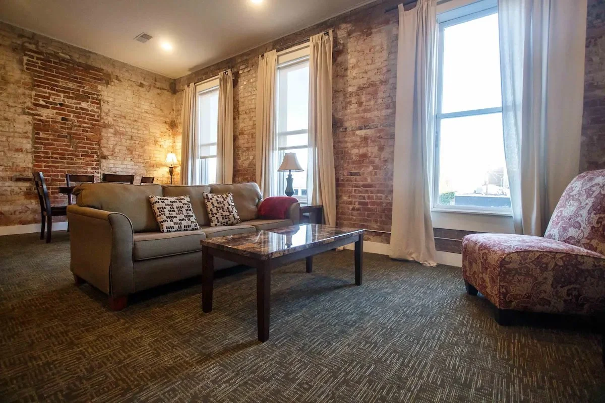 Living room with exposed brick walls, three tall windows with cream curtains, a beige sofa with patterned pillows, a patterned armchair, a wooden coffee table, and two table lamps.