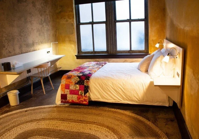 A cozy bedroom with a white bed, colorful quilt, large window, and a small desk with a chair.