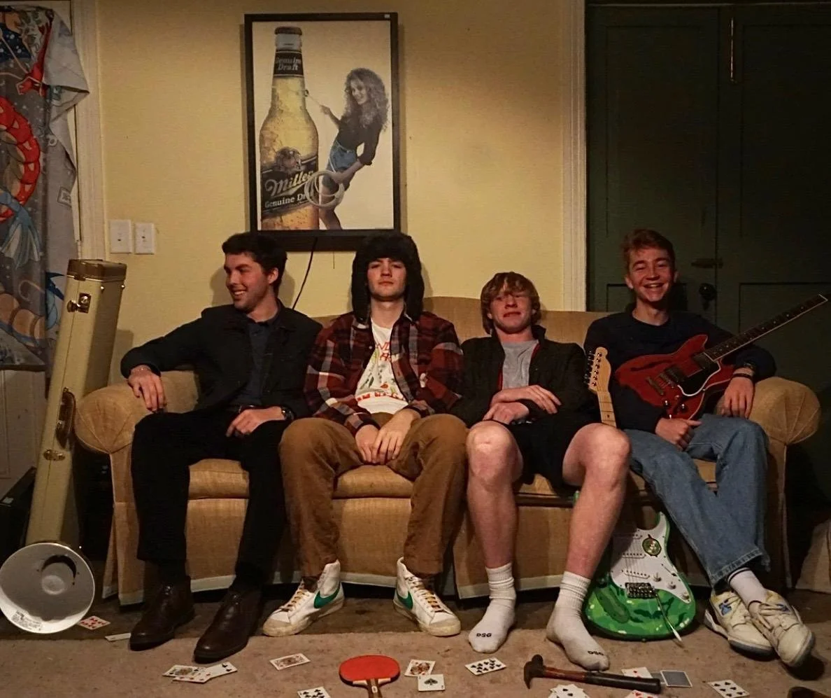 Four young men sitting on a beige couch in a living room, with playing cards scattered on the floor. The first man on the left wears a black jacket and has a smile. The second man has dark hair, a checkered shirt, and is looking straight at the camer