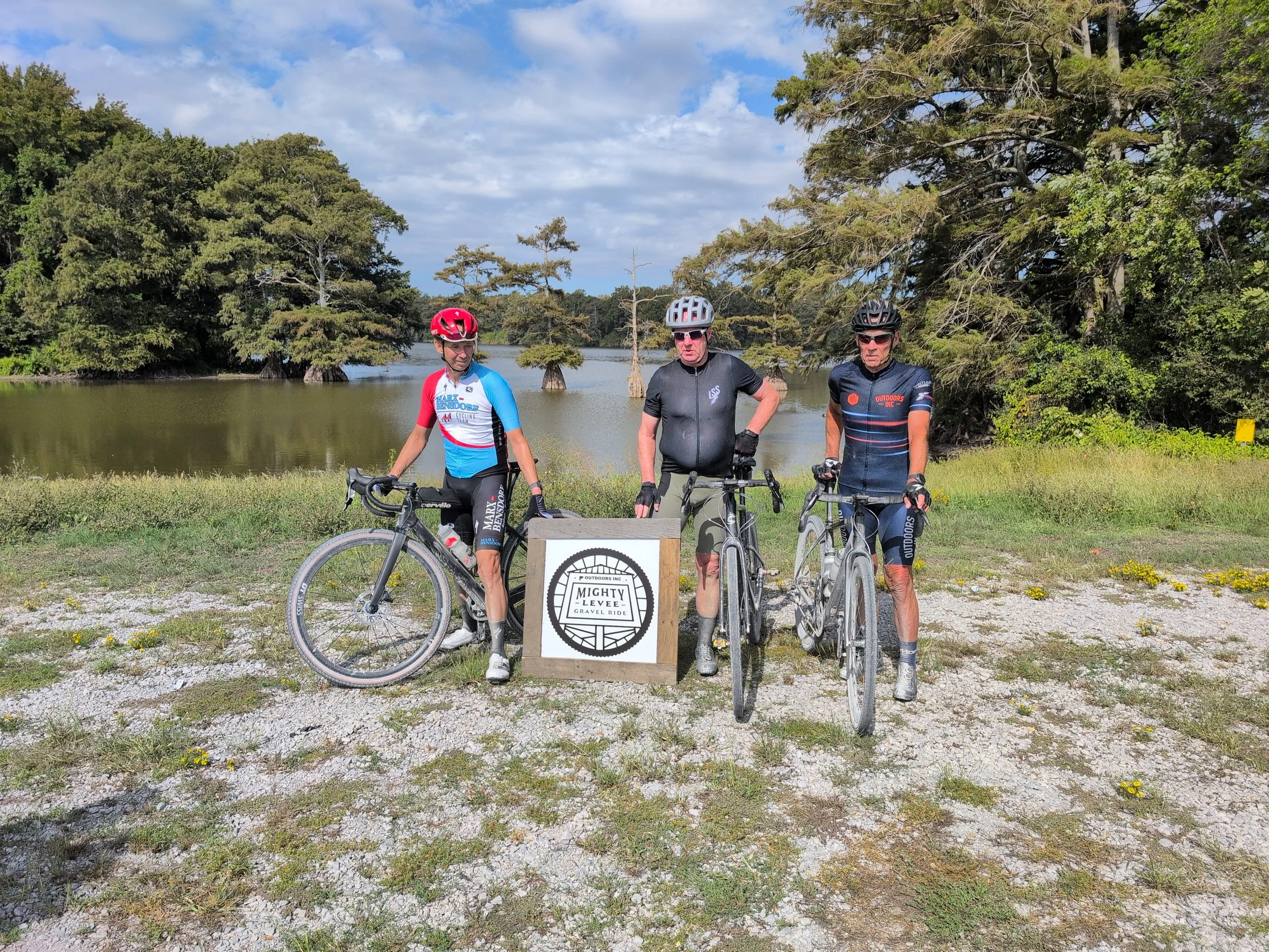 Three cyclists posing with their bikes outdoors near a lake with trees in the background. They are wearing cycling gear and helmets, standing behind a sign that reads 'Mighty Levee Gravel Ride.' The sky is partly cloudy.