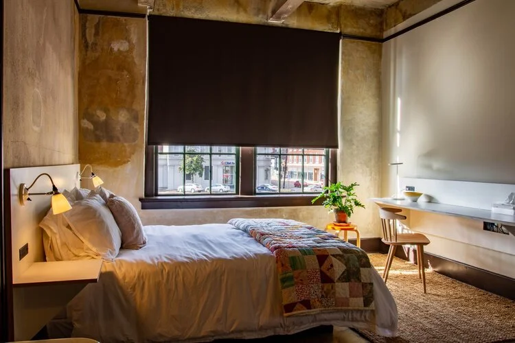 A cozy hotel bedroom with a bed, bedside lamps, a window with city view, a potted plant, and a desk with a chair.