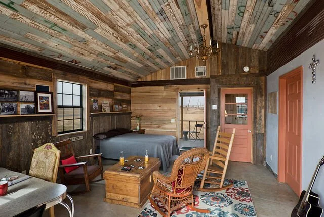 Interior of a rustic cabin bedroom with wooden walls and ceiling, bed with dark bedding, armchair, rocking chair, wooden chest, and door leading outside. Natural light from window and door, decorative cross on wall, and a small rug on the floor.