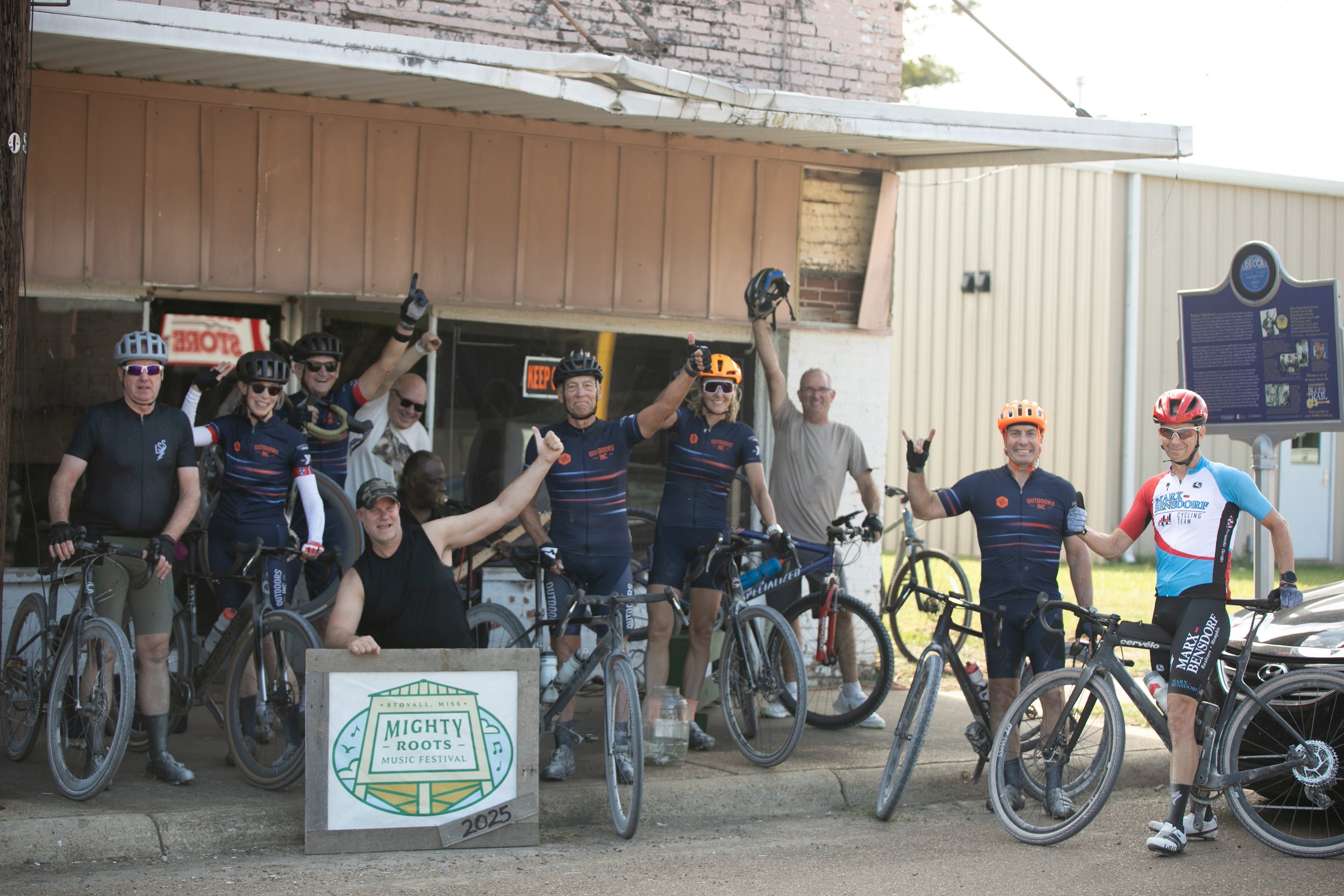 Group of cyclists and supporters posing with their bicycles outside a building at the Mighty Roots Music Festival, Stovall, Mississippi, 2023.