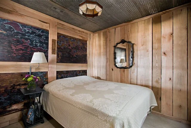 A cozy bedroom with wood-paneled walls, a white bedspread, a small bedside table with a lamp and flowers, a mirror on the wall, and a dark ceiling with a light fixture.