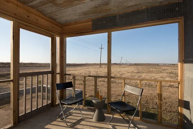View from the porch of an unfinished wooden structure overlooking a flat, open field with power lines in the distance on a clear day.