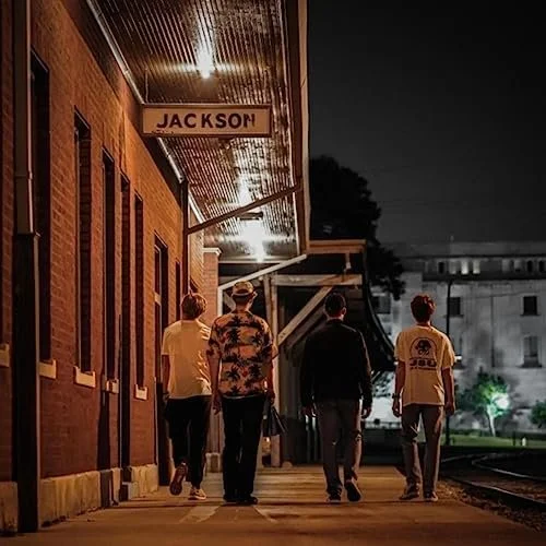 Four people walking along a train station platform at night, with a brick building and a sign that says 'Jackson' overhead.