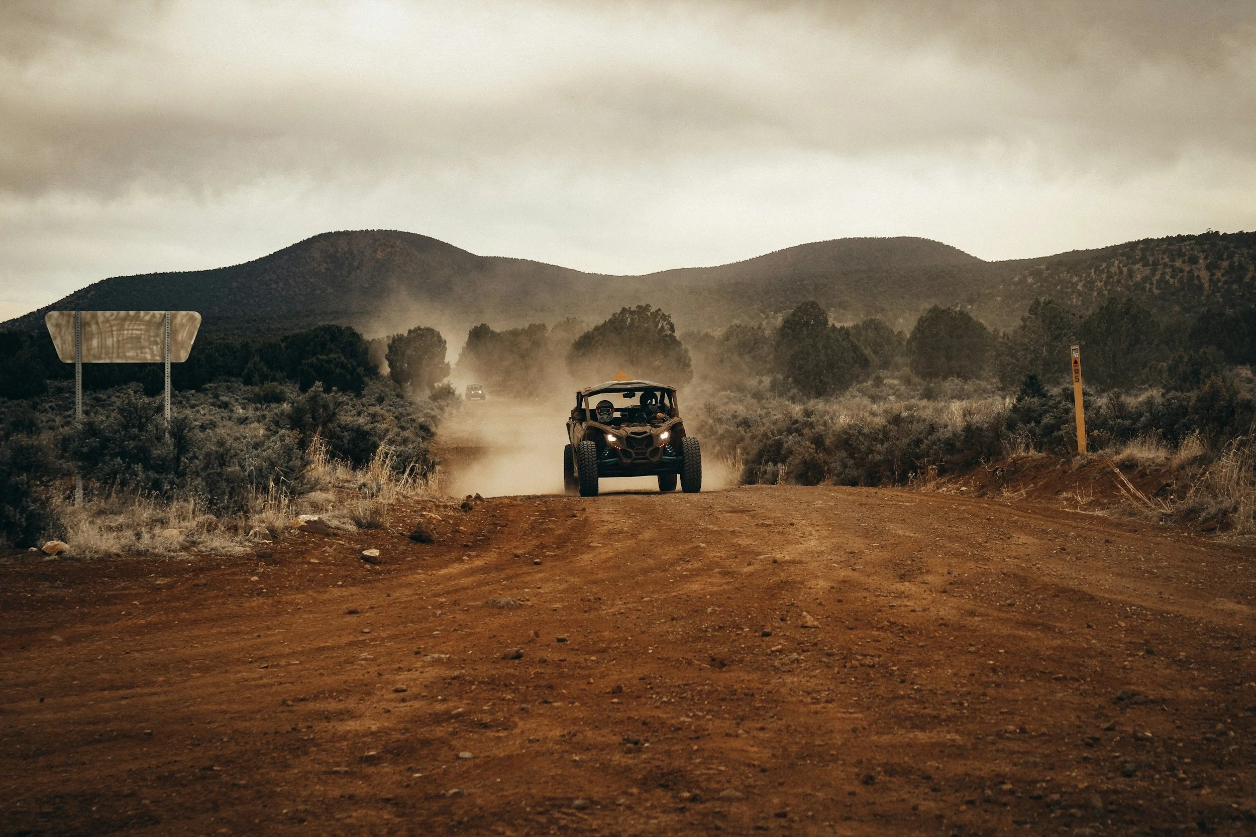 An off-road utility vehicle driving on a dirt trail through a semi-arid landscape with mountains and cloudy sky in the background.