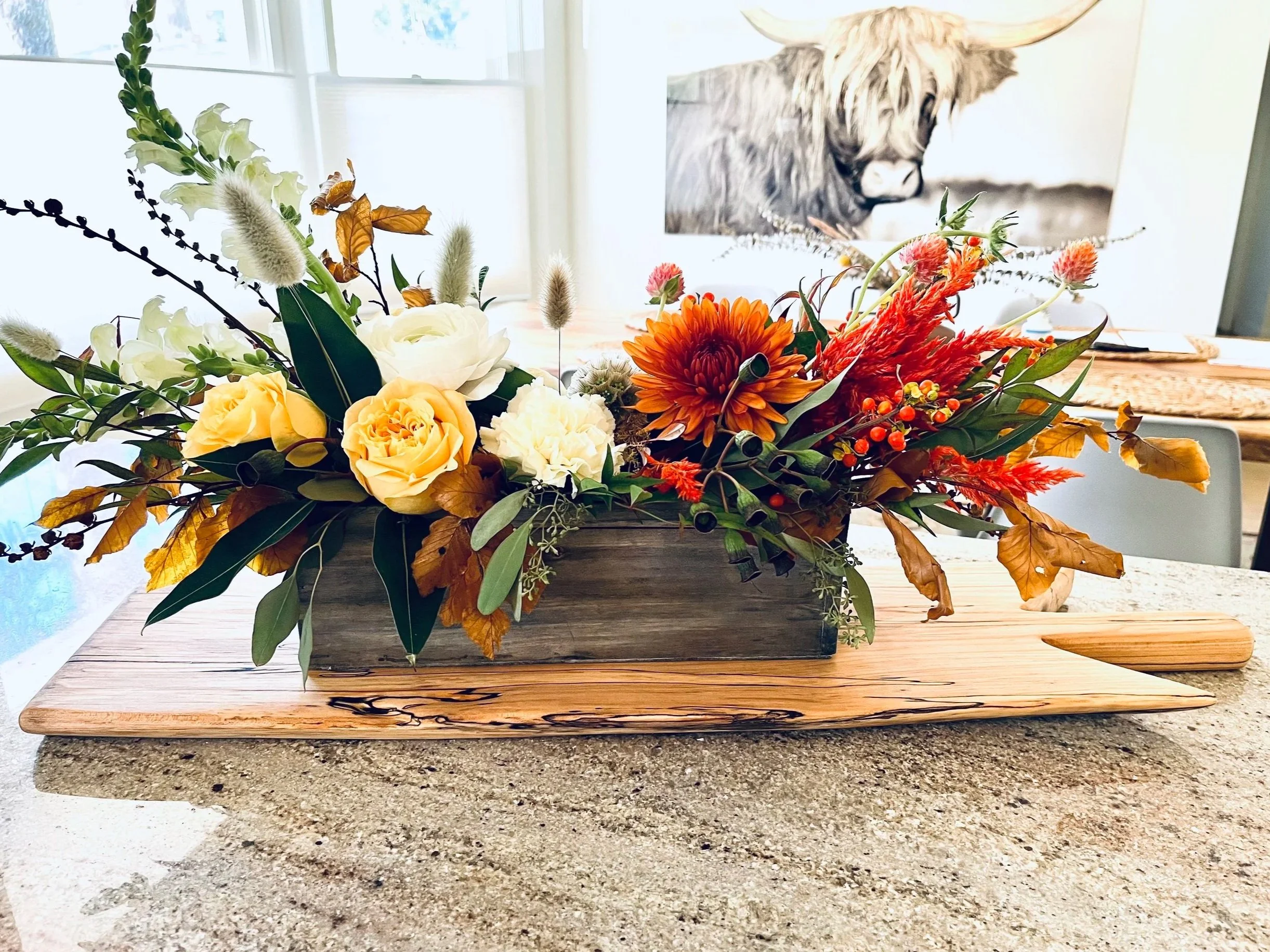 A floral arrangement with yellow and white flowers, green foliage, and orange accents in a wooden container on a wooden tray.