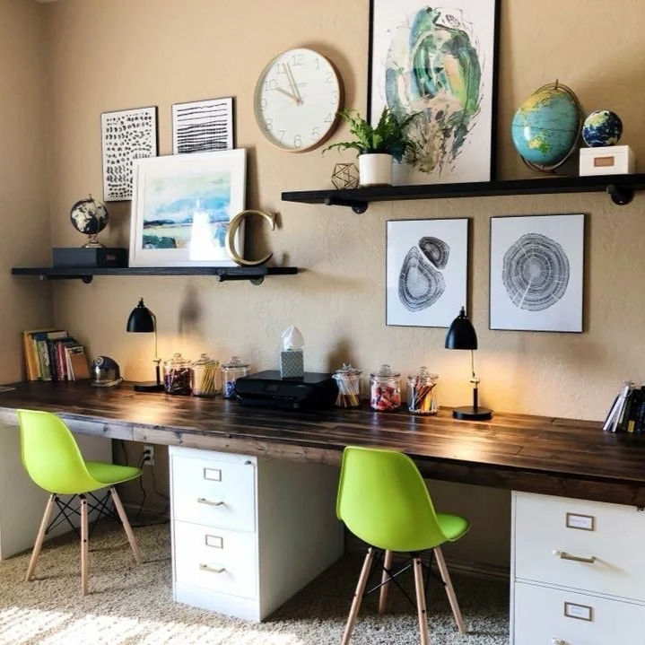 Home office with a long wooden desk, two bright green chairs, and a beige wall decorated with framed artwork, shelves, and a wall clock. The desk has jars with supplies, a lamp, and office equipment.