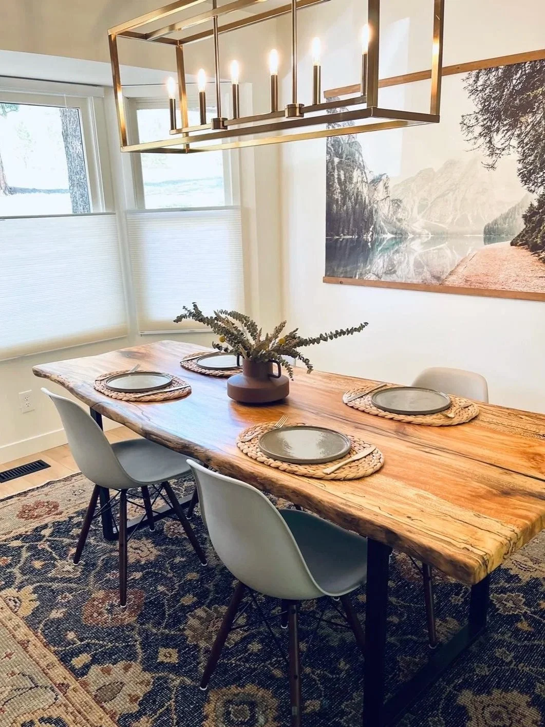 A dining room with a large rustic wooden table set with four round placemats, a brown vase with greenery as a centerpiece, and four white modern chairs. A rectangular chandelier with candle-like lights hangs above the table. There is a large landscap