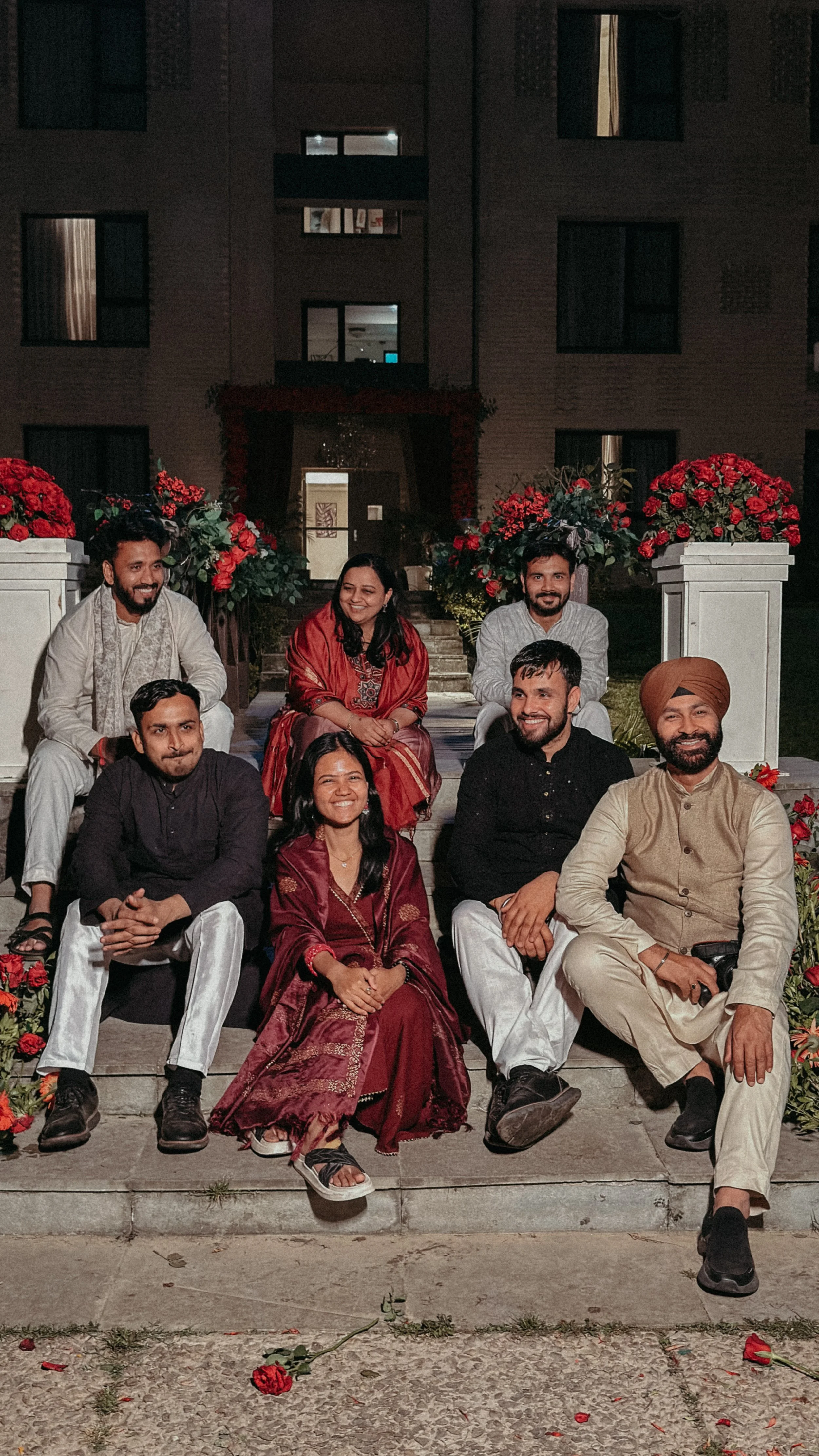 Group of seven people celebrating at night in traditional Indian attire, seated and standing on steps in front of a decorated entrance with red flowers, in an outdoor setting.