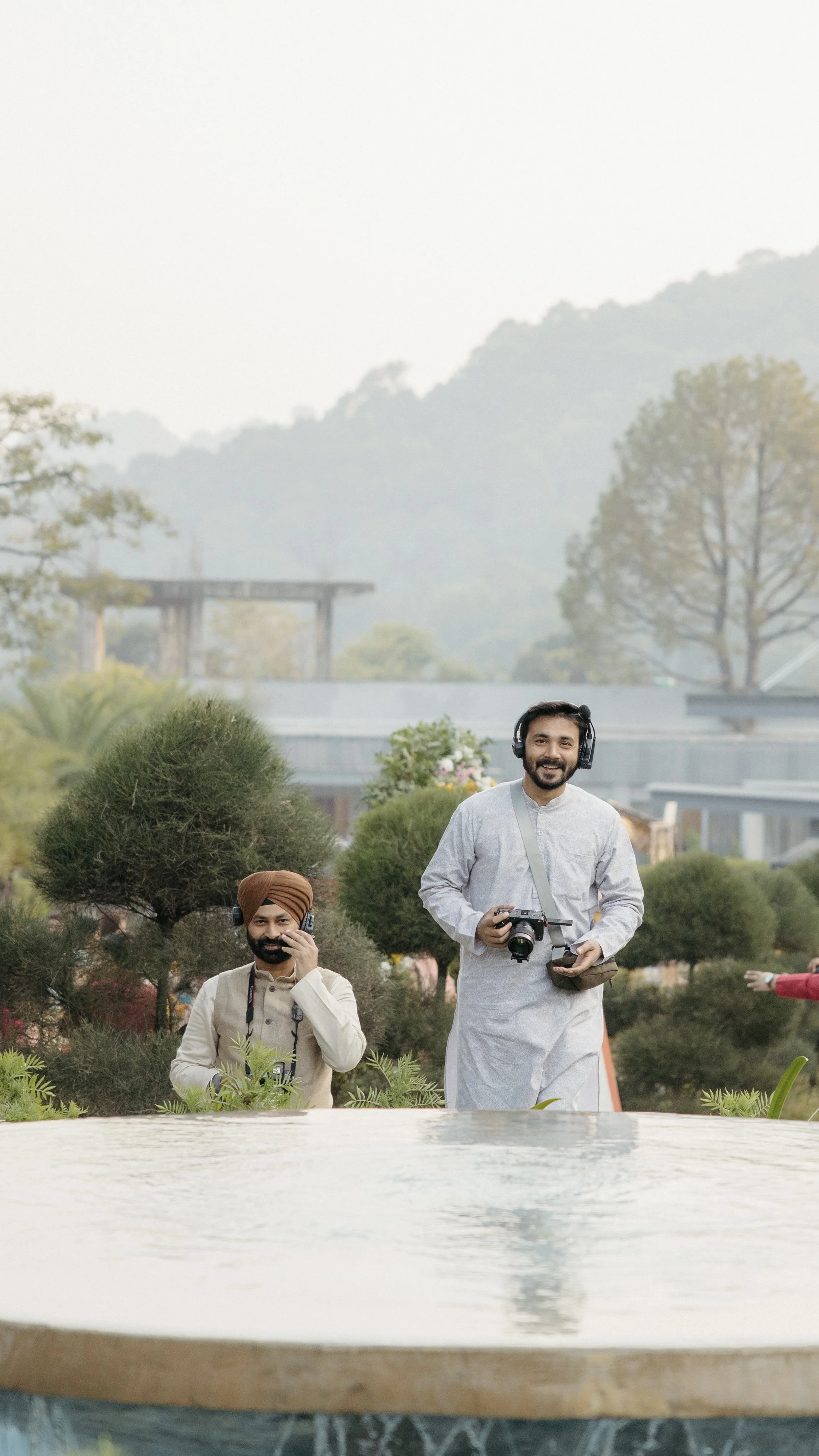 Two men wearing headphones and holding cameras, smiling in a lush garden with trees and shrubs, with hills and a building in the background.