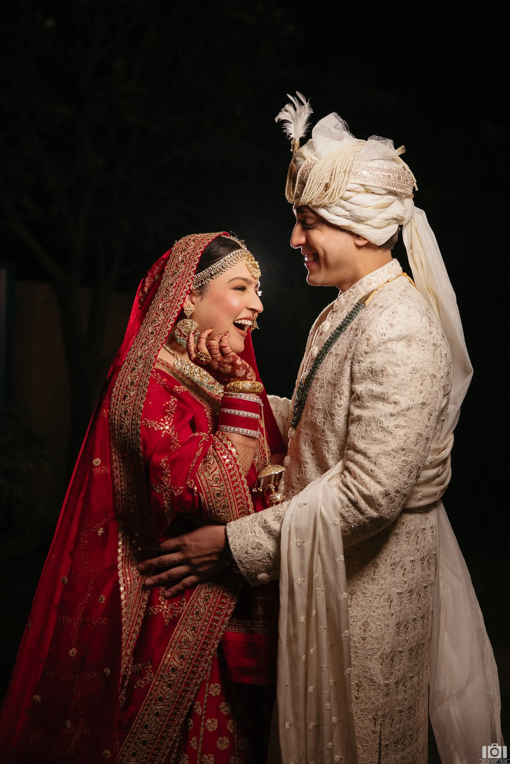 A happy bride and groom in traditional Indian wedding attire, smiling and embracing each other at night, with dark trees in the background.