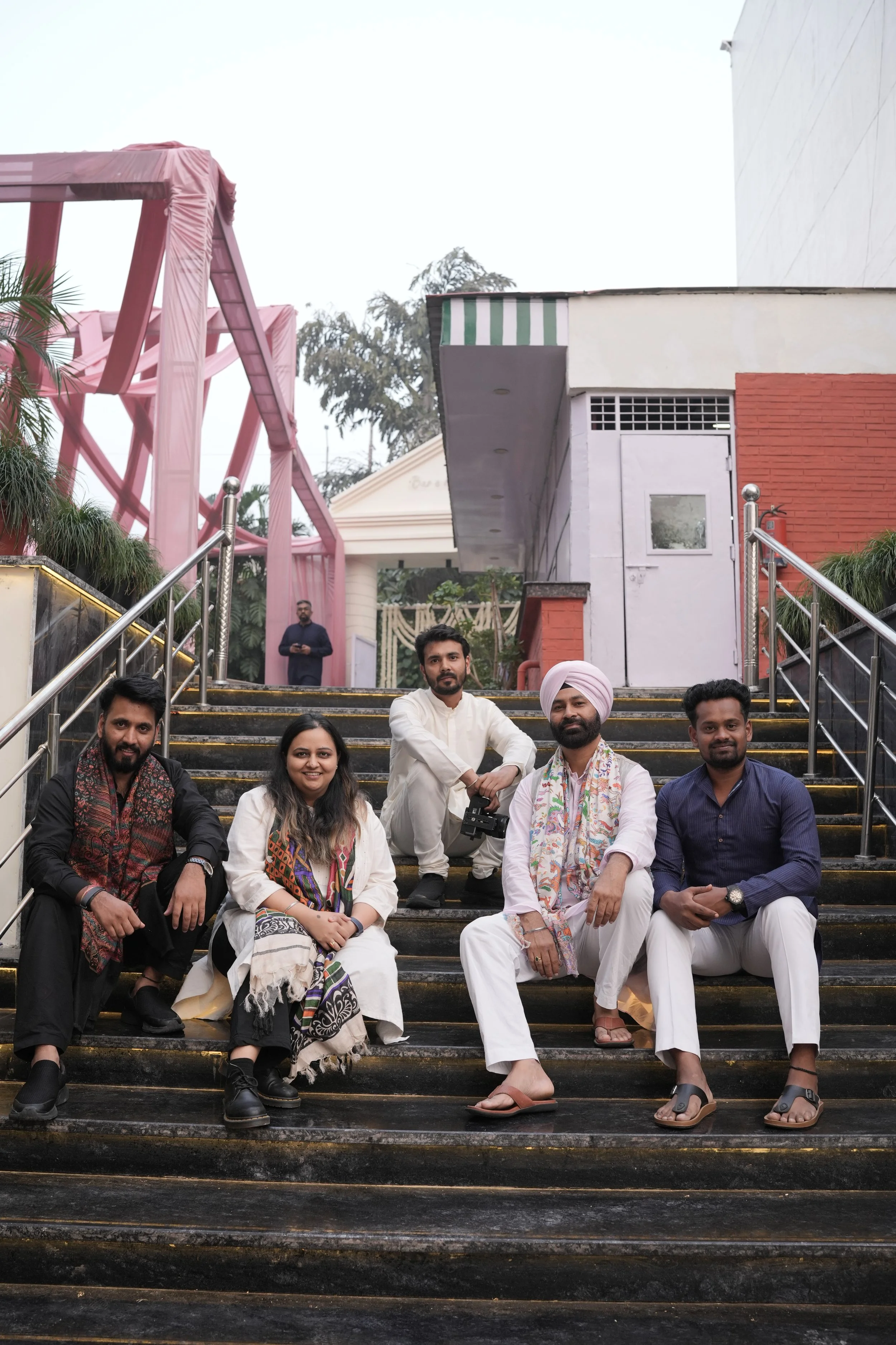 Group of seven Indian people sitting and posing on a staircase outside a building, with one person standing in the background. The group includes men and women dressed in traditional and modern clothing.
