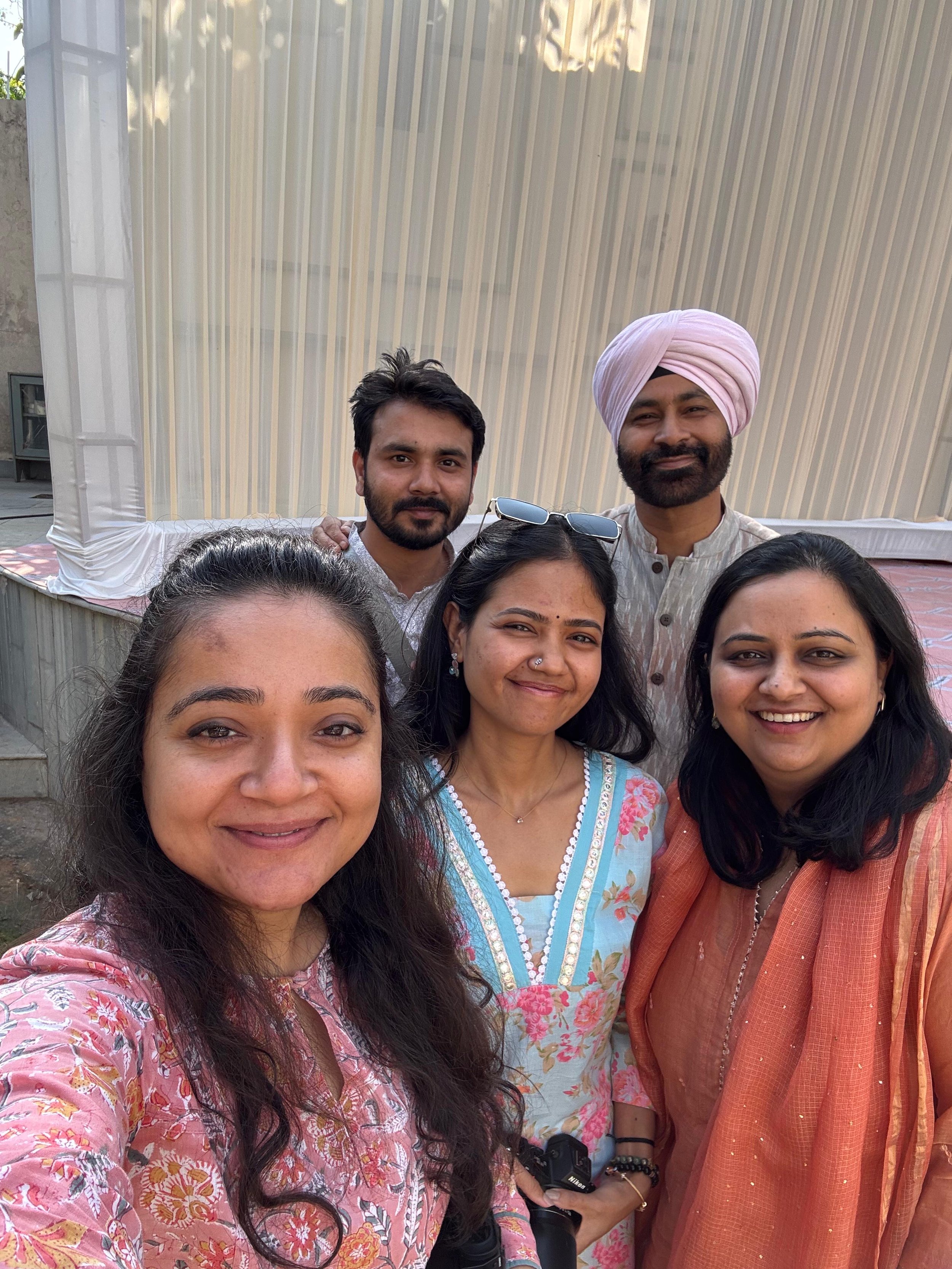 Group of six friends taking a selfie together outdoors, some wearing traditional Indian attire and one holding a Nikon camera, with a light-colored fabric structure in the background.