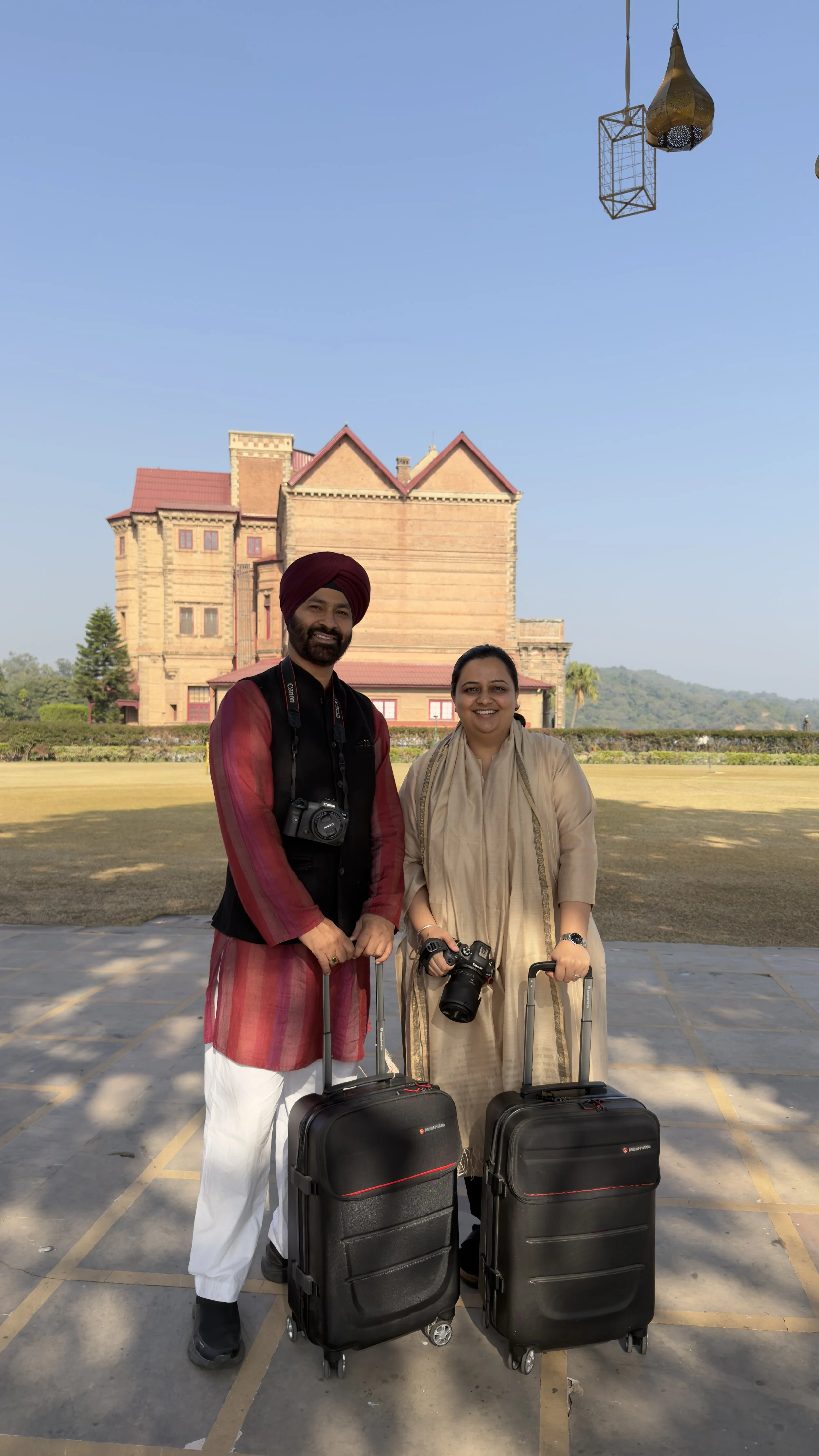 A man and woman standing outdoors with rolling luggage, smiling, in front of a historic building with a red roof, under a clear blue sky.