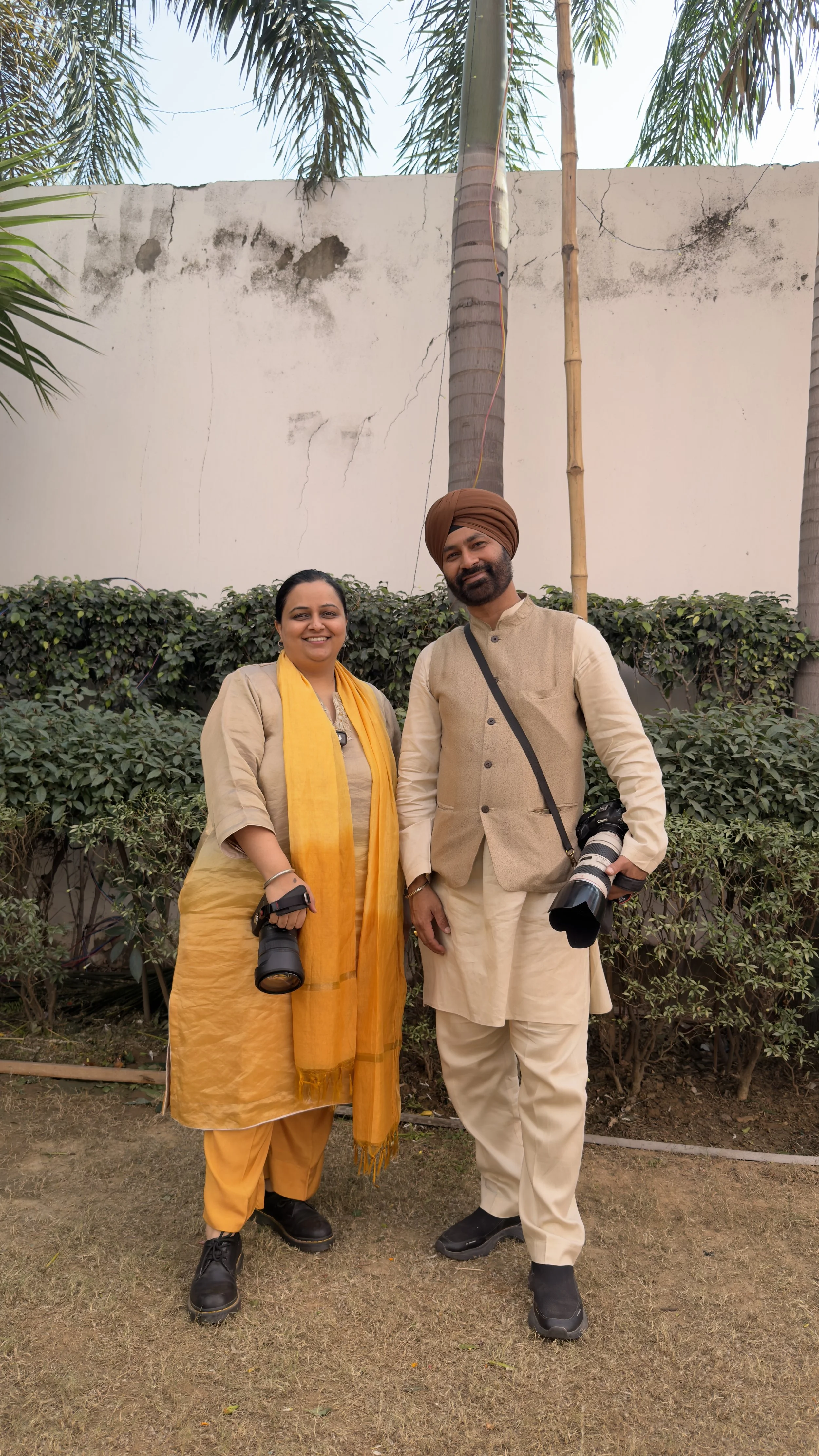A man and woman standing outdoors in front of a white wall with palm trees. The woman is wearing a yellow scarf and traditional Indian outfit, holding a camera. The man is dressed in cream-colored traditional Indian attire, wearing a turban, and hold