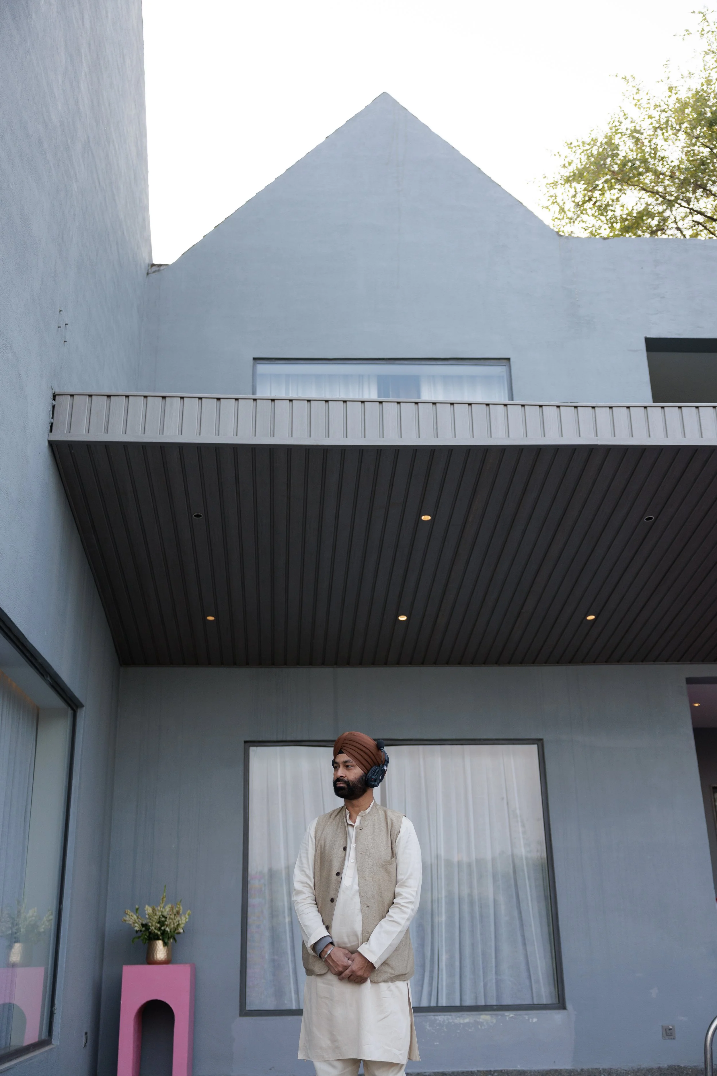A man wearing traditional Indian attire with a beige vest over a white kurta and a matching dhoti stands with folded hands in front of a modern building with large windows, a balcony, and a small pink table with a vase of flowers, with a person weari