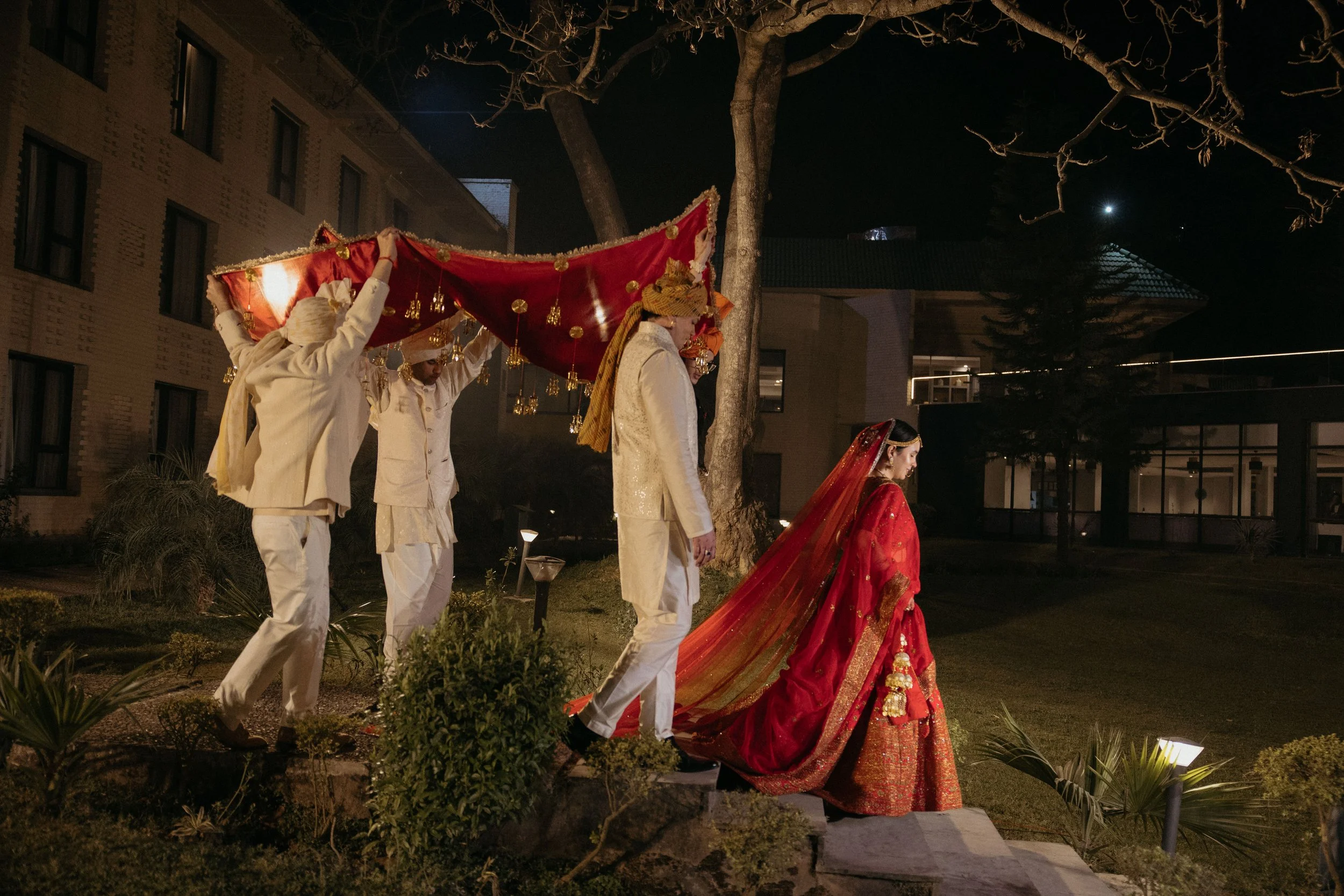 People participating in a traditional Indian wedding ceremony at night, with the bride dressed in a red and gold bridal outfit and the groom in white, under a decorative canopy held by others.
