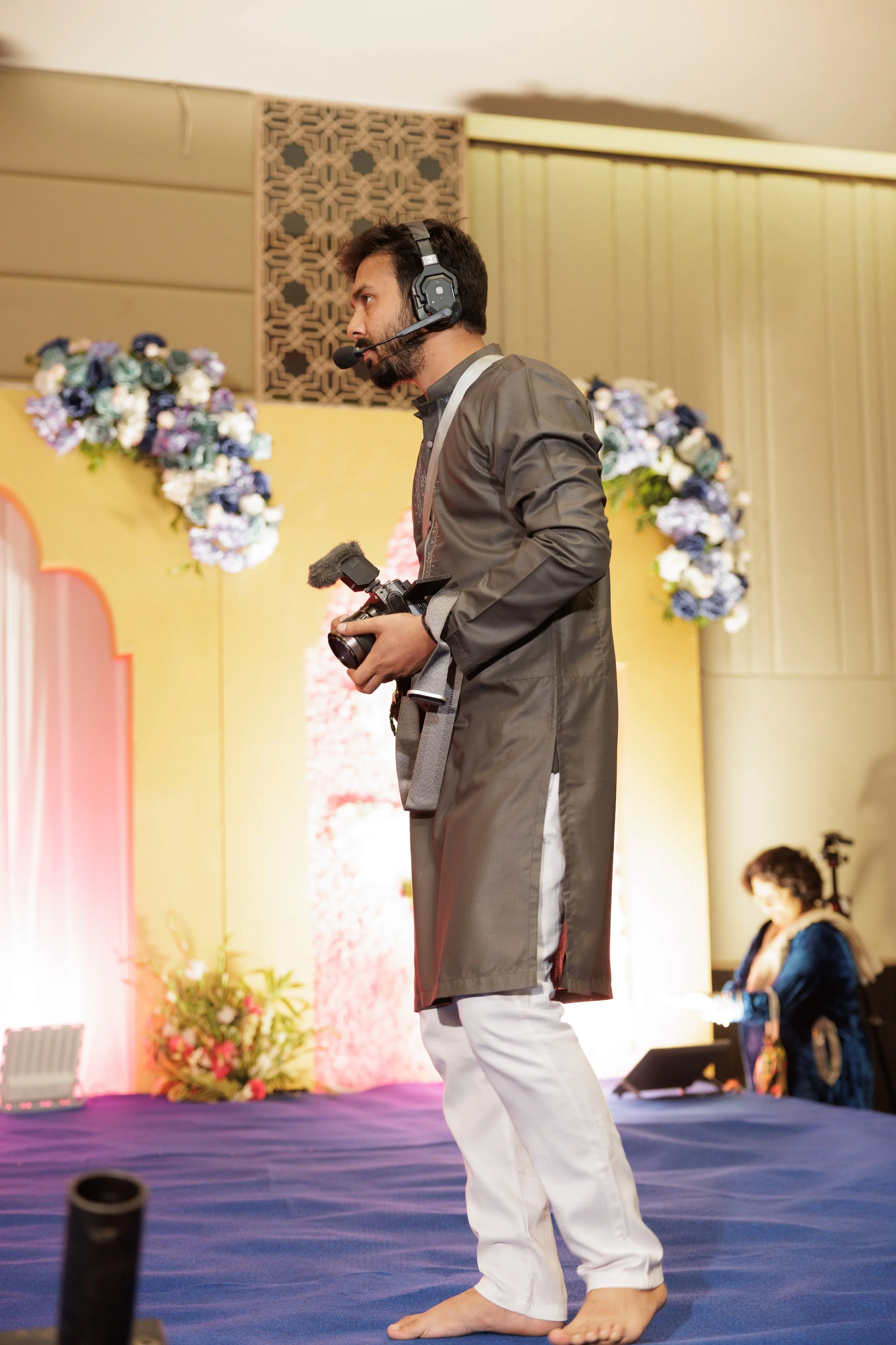 A man wearing a headset with a microphone, holding a camera, standing on a stage decorated with flowers, with a woman seated in the background.