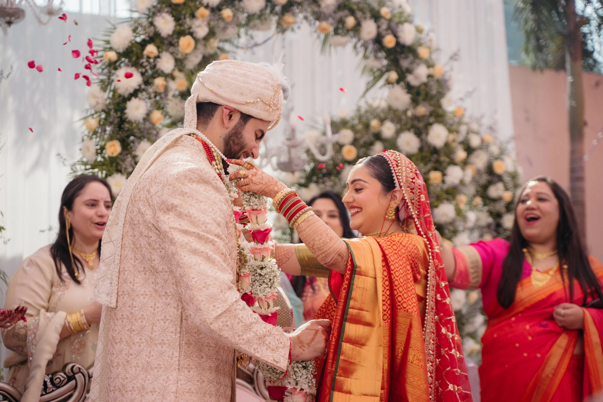 A couple in traditional Indian attire participating in a wedding ceremony surrounded by women in colorful dresses, with a floral backdrop and petals falling.