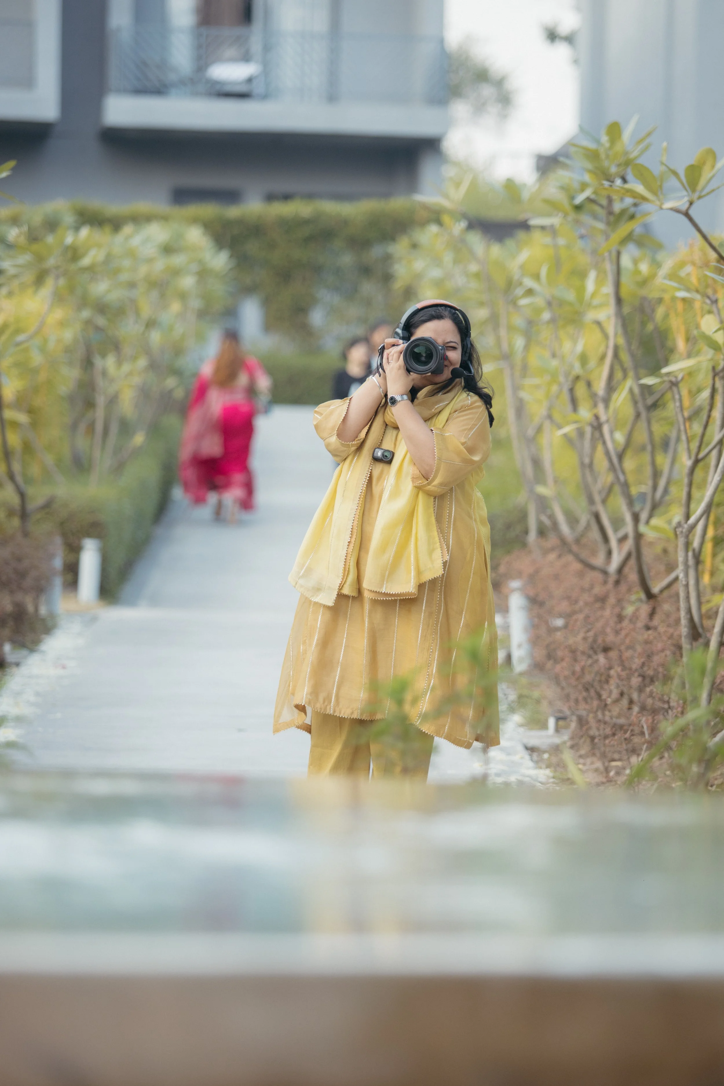 A woman in a yellow outfit is taking a photo with a camera, standing on a pathway surrounded by greenery, with other people in the background.